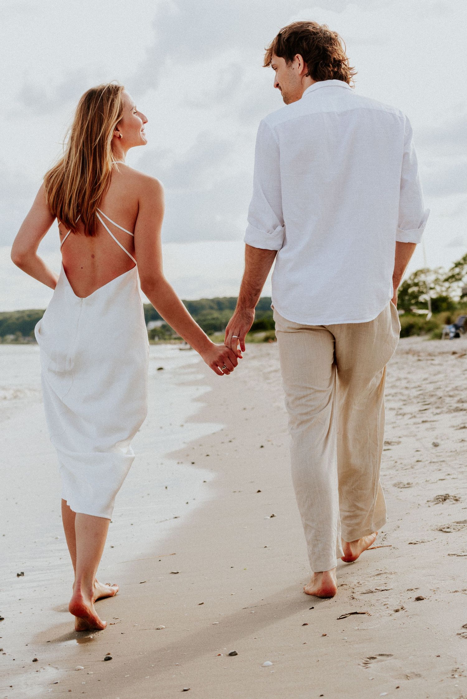 A couple walking hand in hand along a sandy beach wearing white and beige casual attire.