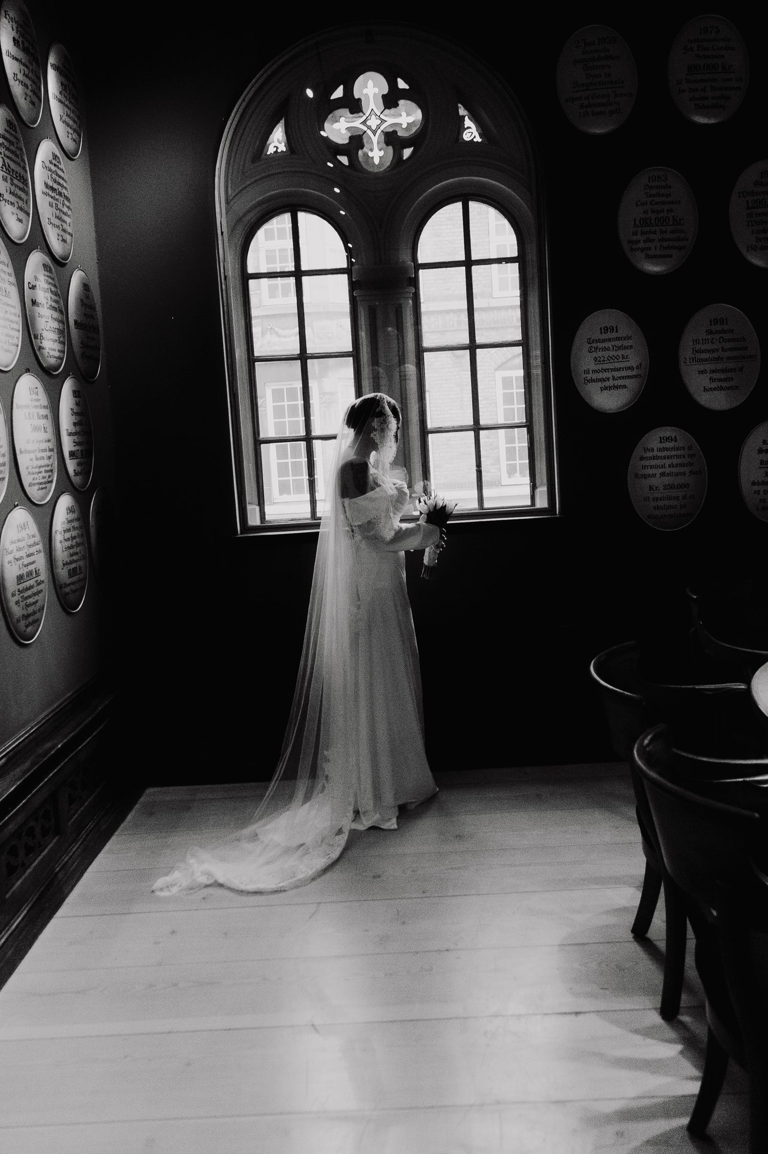 Elegant black and white photo of a bride in flowing dress and veil standing by arched gothic windows.