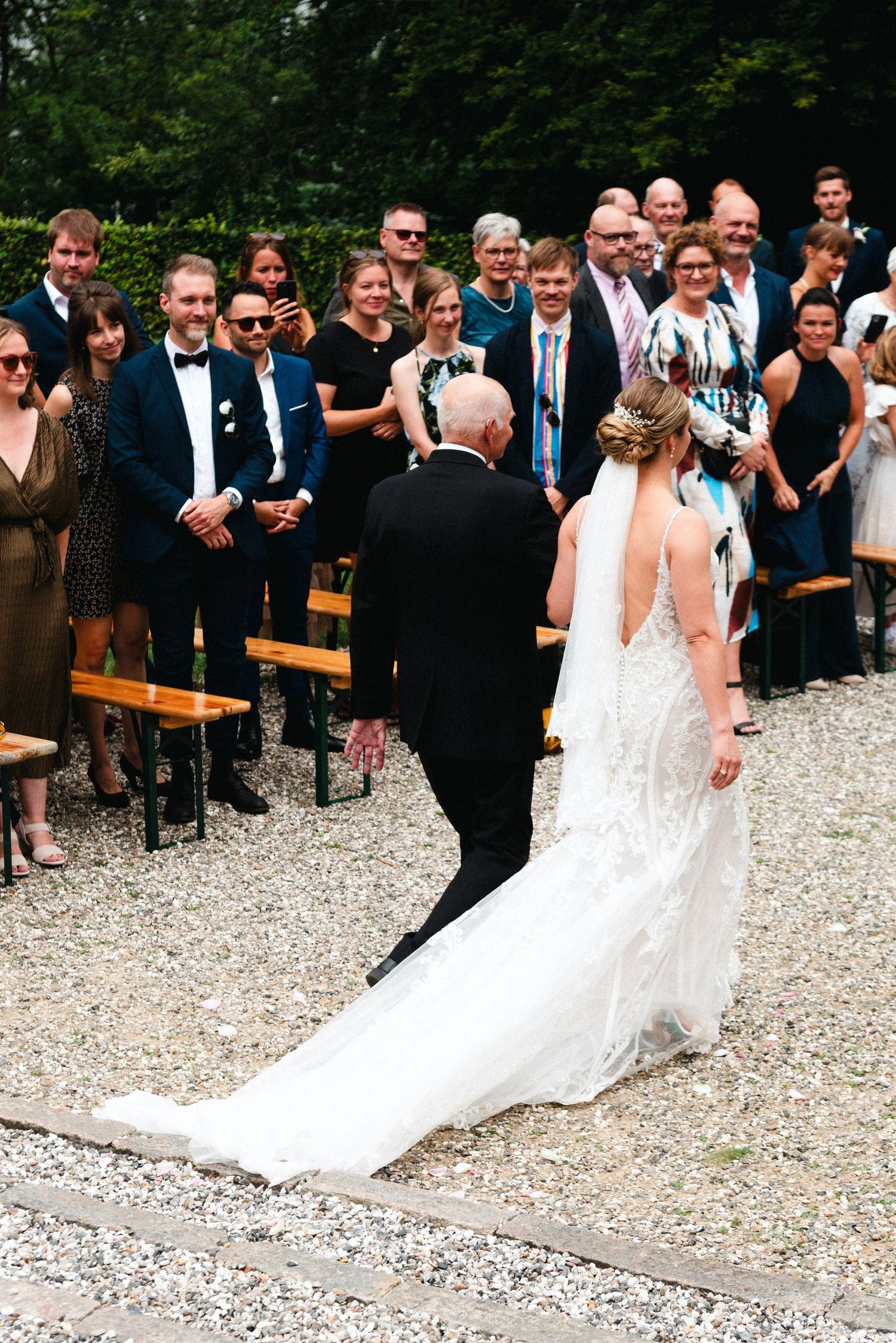 Wedding ceremony procession on gravel path with guests watching from wooden benches.