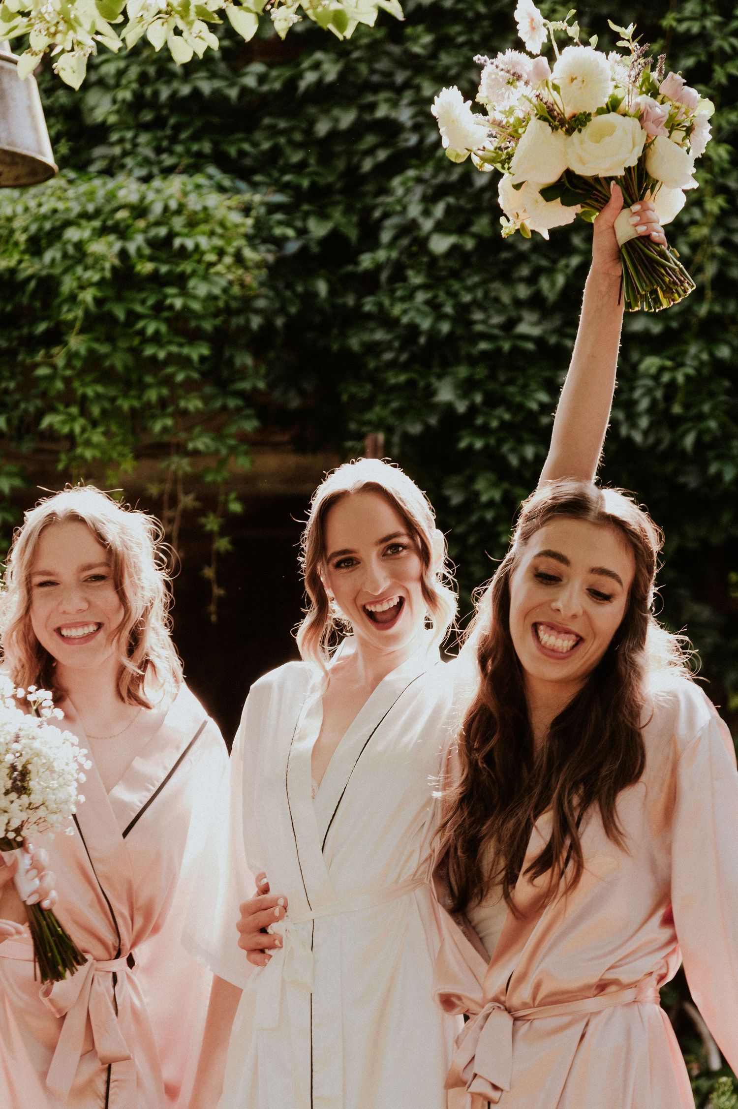 A group of bridesmaids in pink robes laugh and smile together during wedding day preparations outdoors.