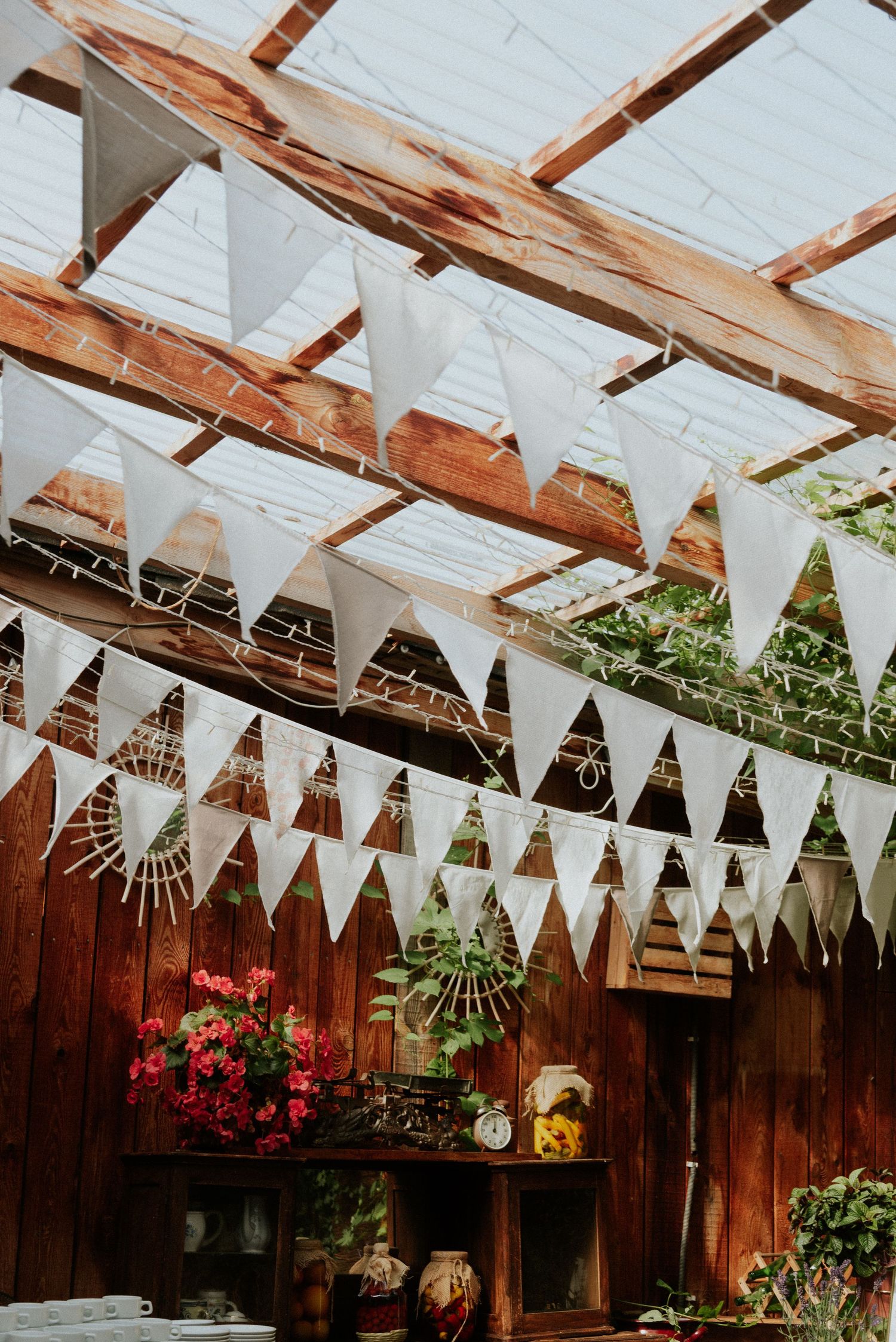 White triangular bunting flags hang from wooden beams in a rustic greenhouse with plants and red flowers.