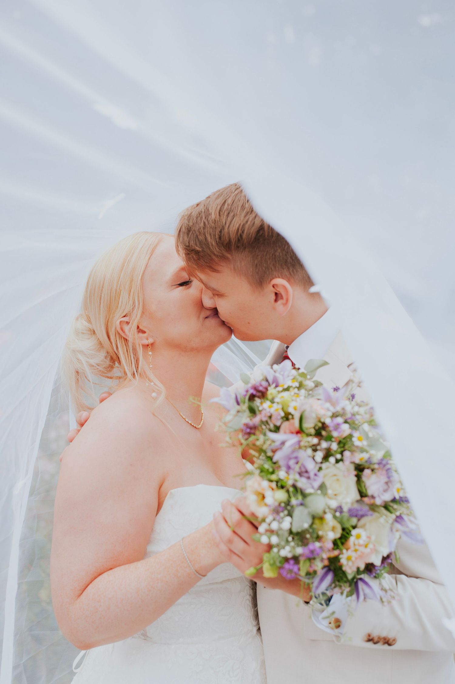 Close-up shot of a romantic wedding kiss with delicate white and purple bouquet.