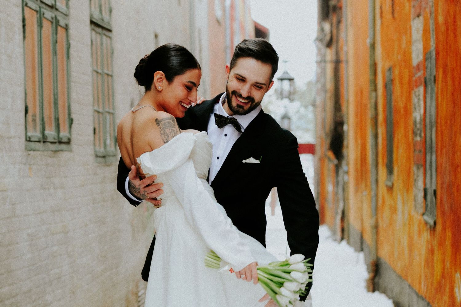 A joyful wedding couple embraces and laughs together in an urban alley with rustic orange and white walls.