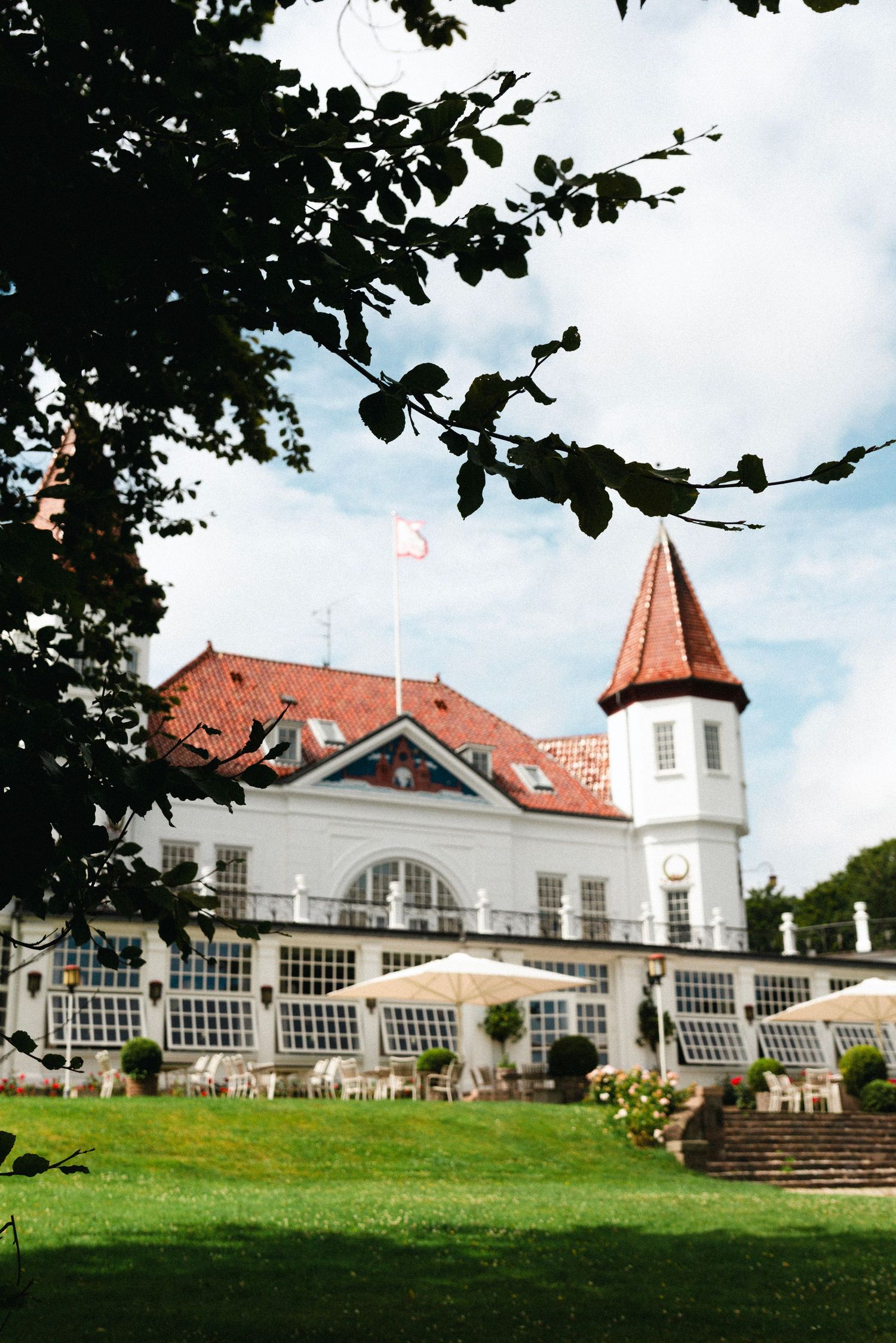 White wedding venue with turrets and arched windows sits on a grassy hill surrounded by trees.