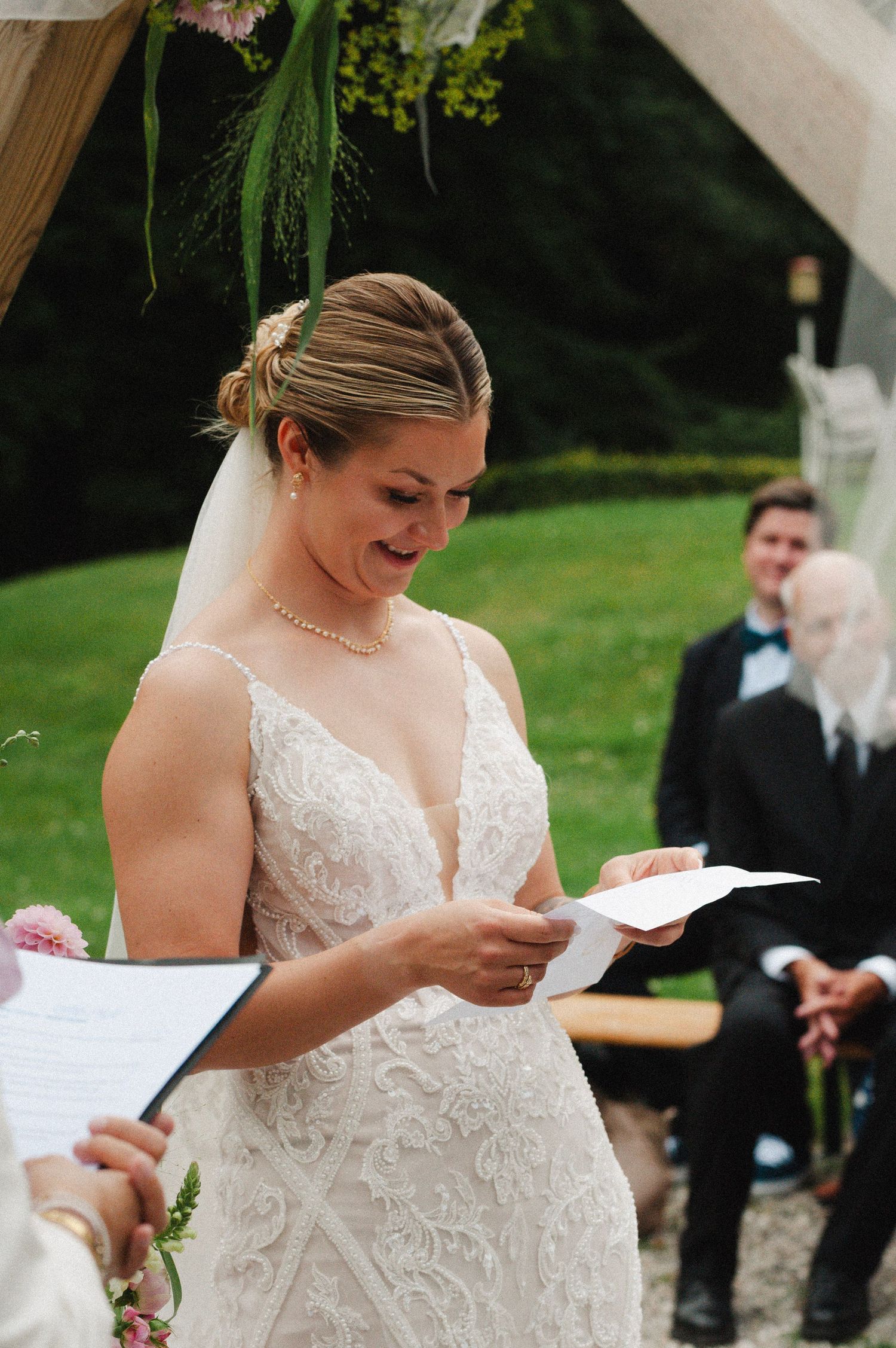 Bride in a white lace wedding dress reads from a paper during an outdoor ceremony.
