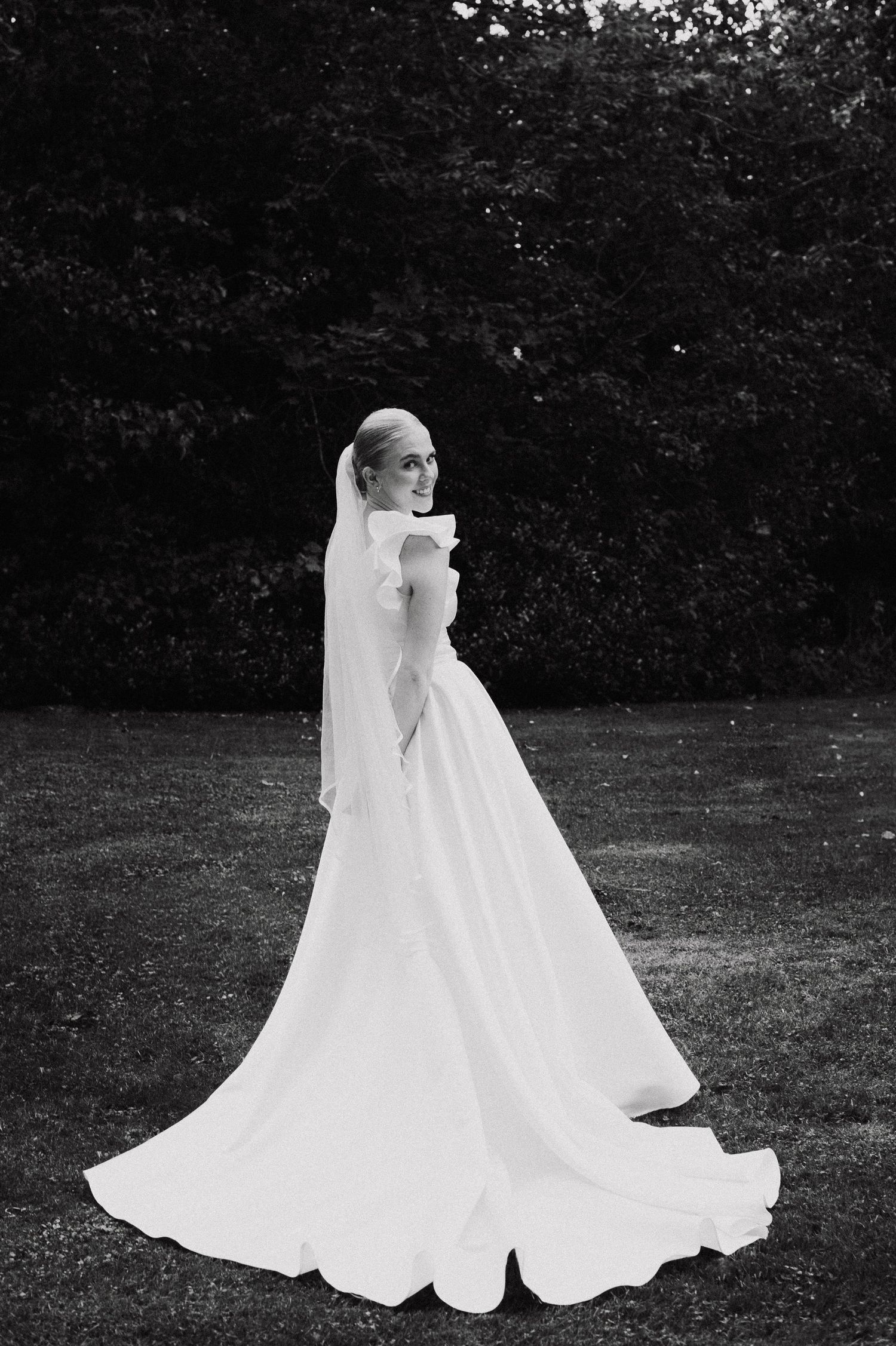 A bride in an elegant white wedding gown with a flowing veil poses in a garden setting, captured in black and white.