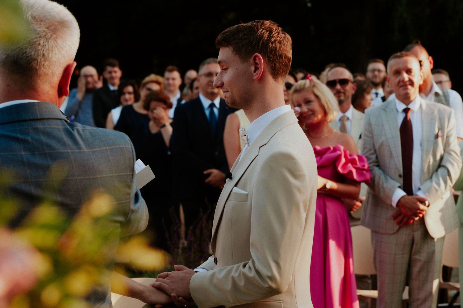 Wedding guests gather outdoors in formal attire with men wearing light colored suits and women in bright dresses.