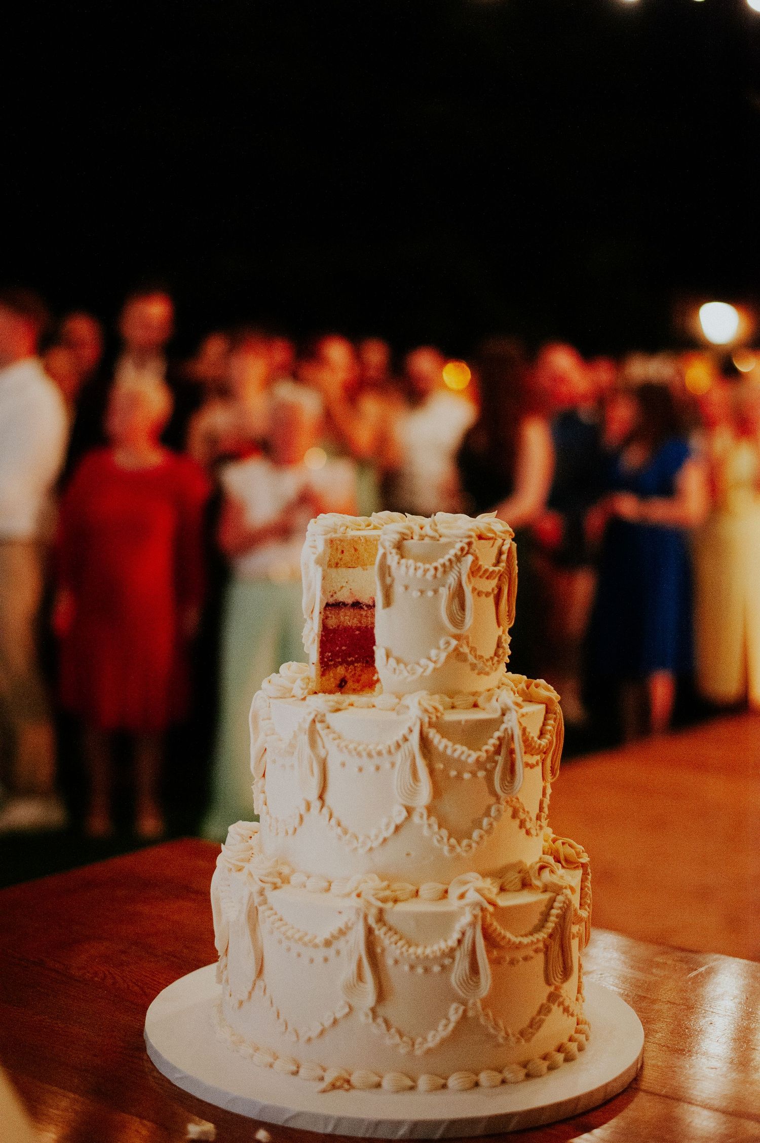 A white tiered wedding cake with delicate pearl piping and scalloped details glows warmly under reception lighting.
