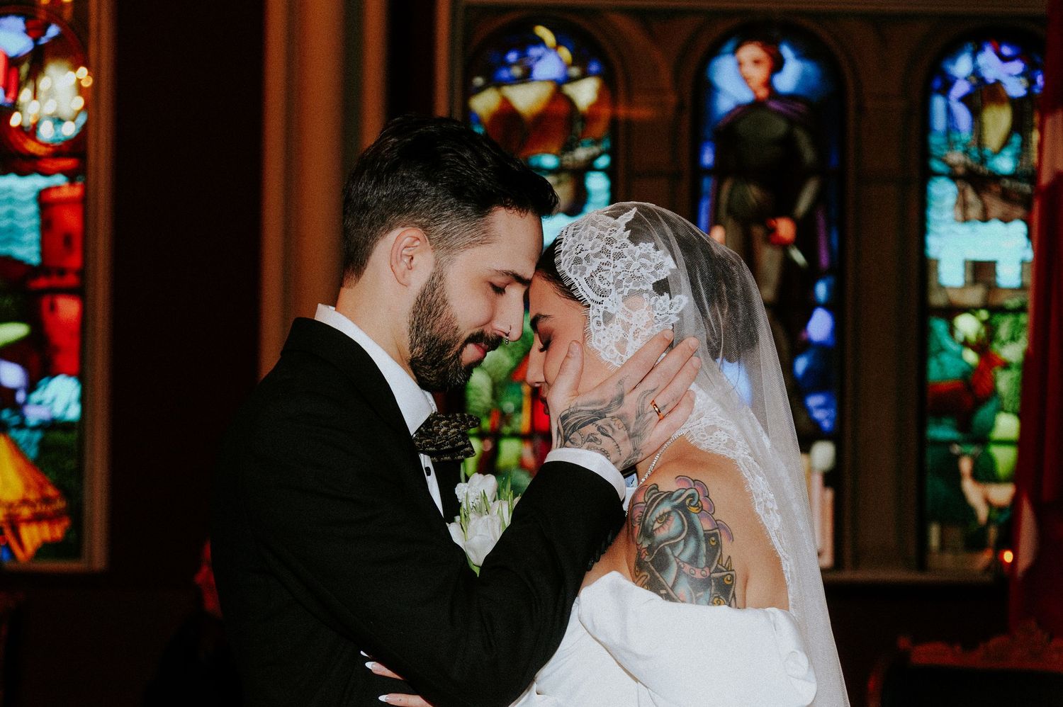 A couple in wedding attire share a romantic kiss during their ceremony in front of stained glass windows.