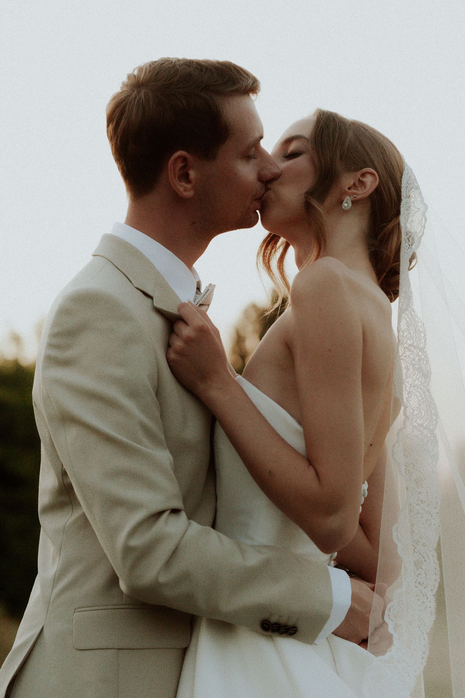 A romantic wedding kiss between a couple, with the bride in white and groom in light beige suit at sunset.