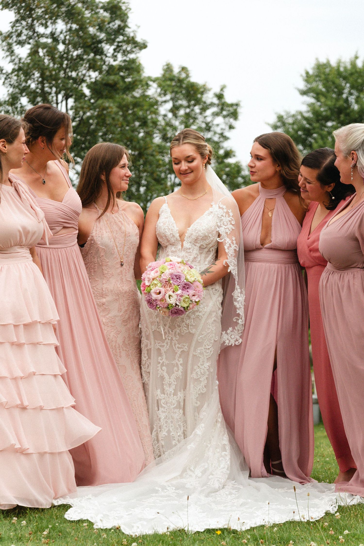 Bridesmaids in blush pink dresses pose with bride in white gown for an outdoor wedding photo.
