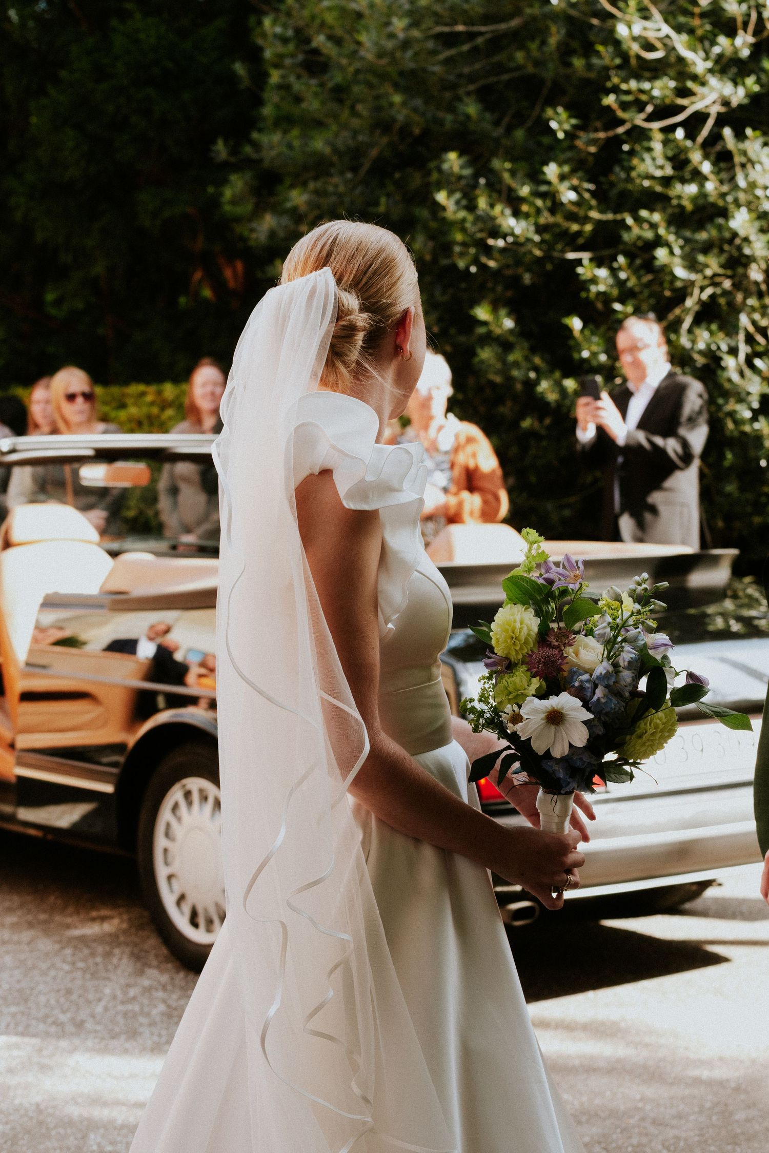 Bride in white dress holding bouquet walks near vintage car on wedding day.