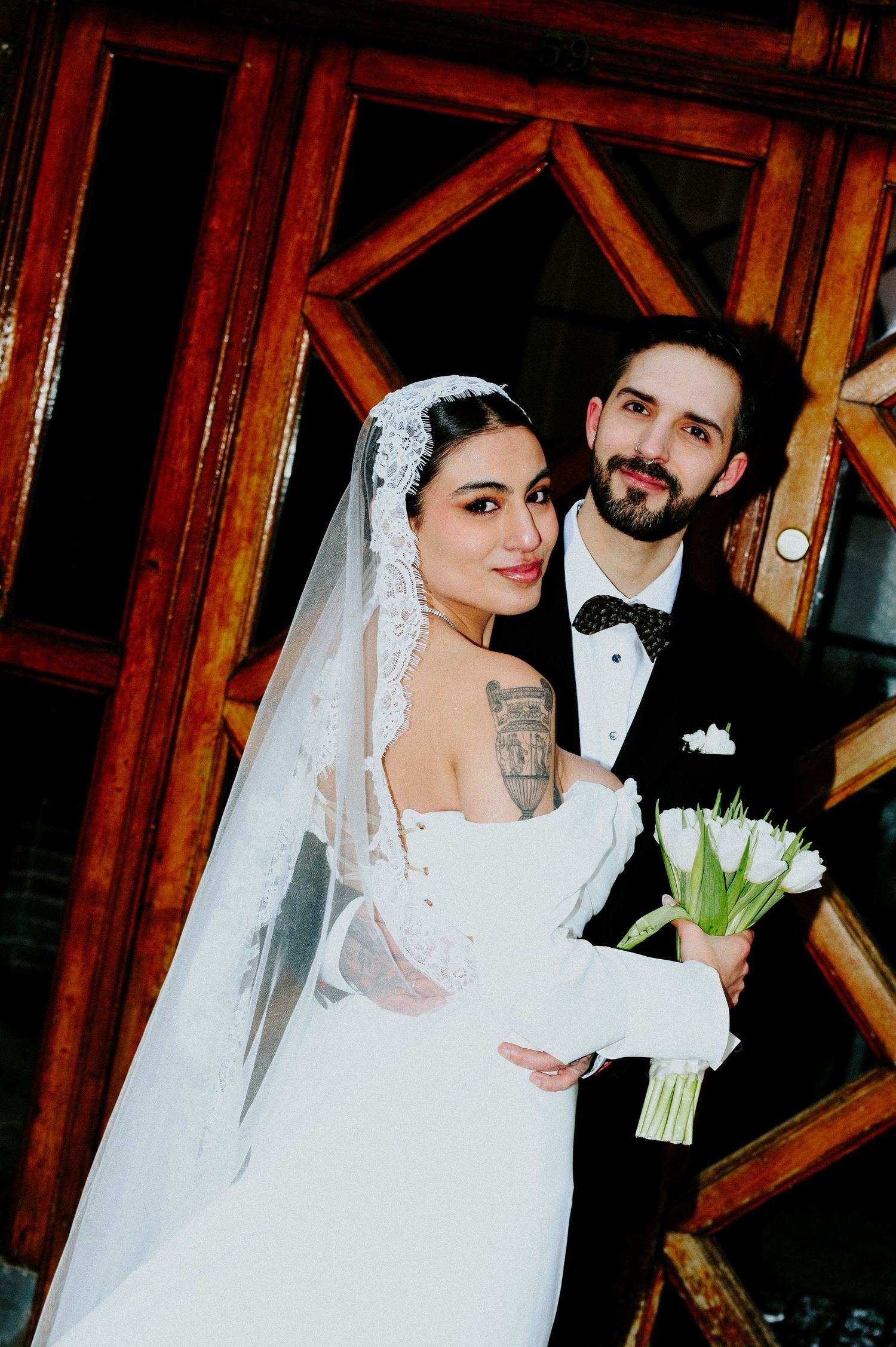A wedding portrait against wooden doors showing a bride in white holding tulips next to her groom in black.