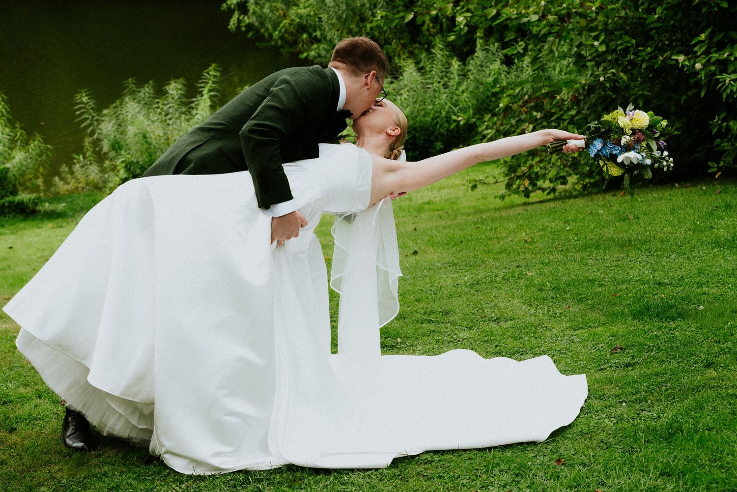 A couple in wedding attire performs a romantic dip pose on a lush green lawn during their outdoor celebration.