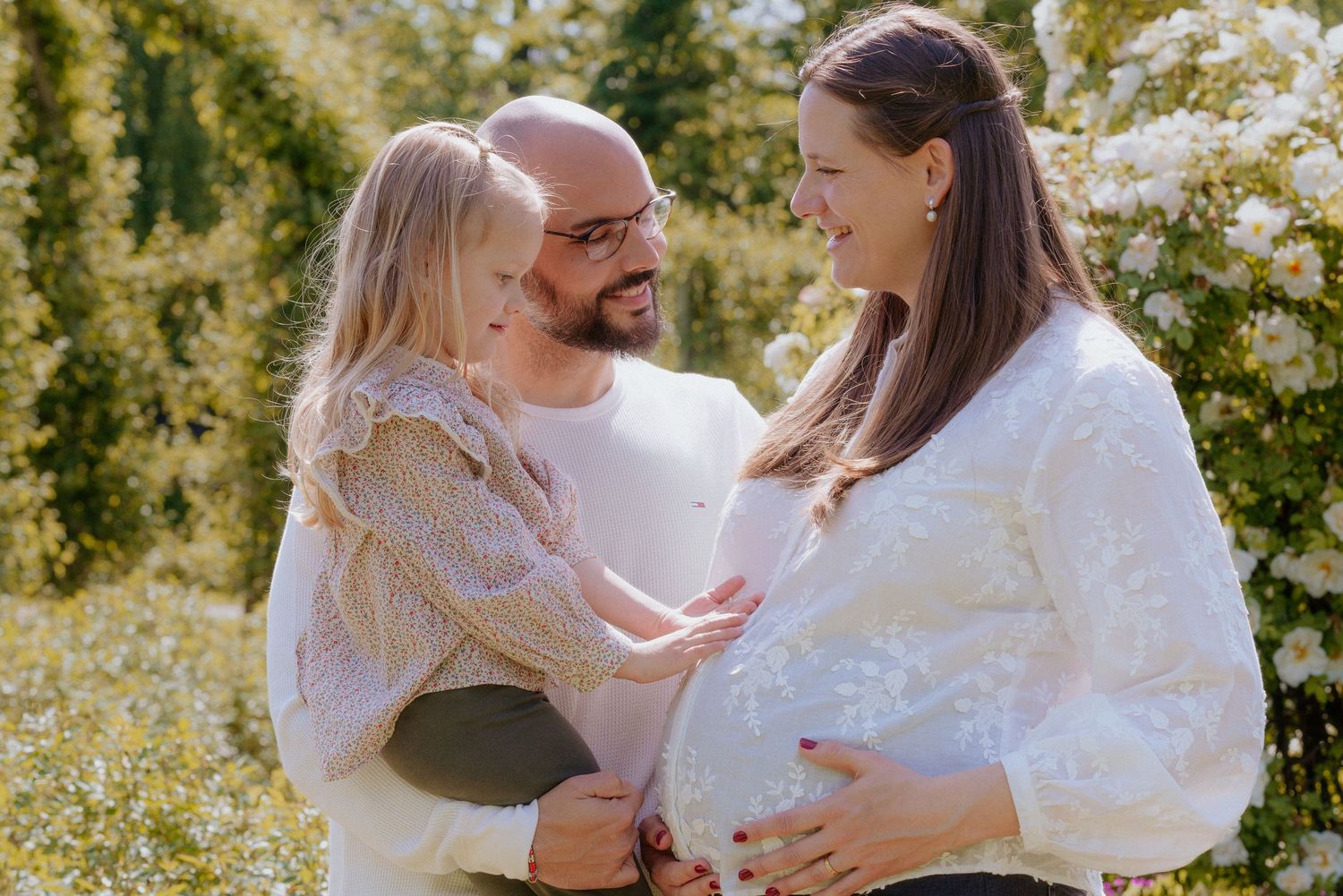 A family in white outfits sits together on grass during an outdoor photo session in a natural setting.