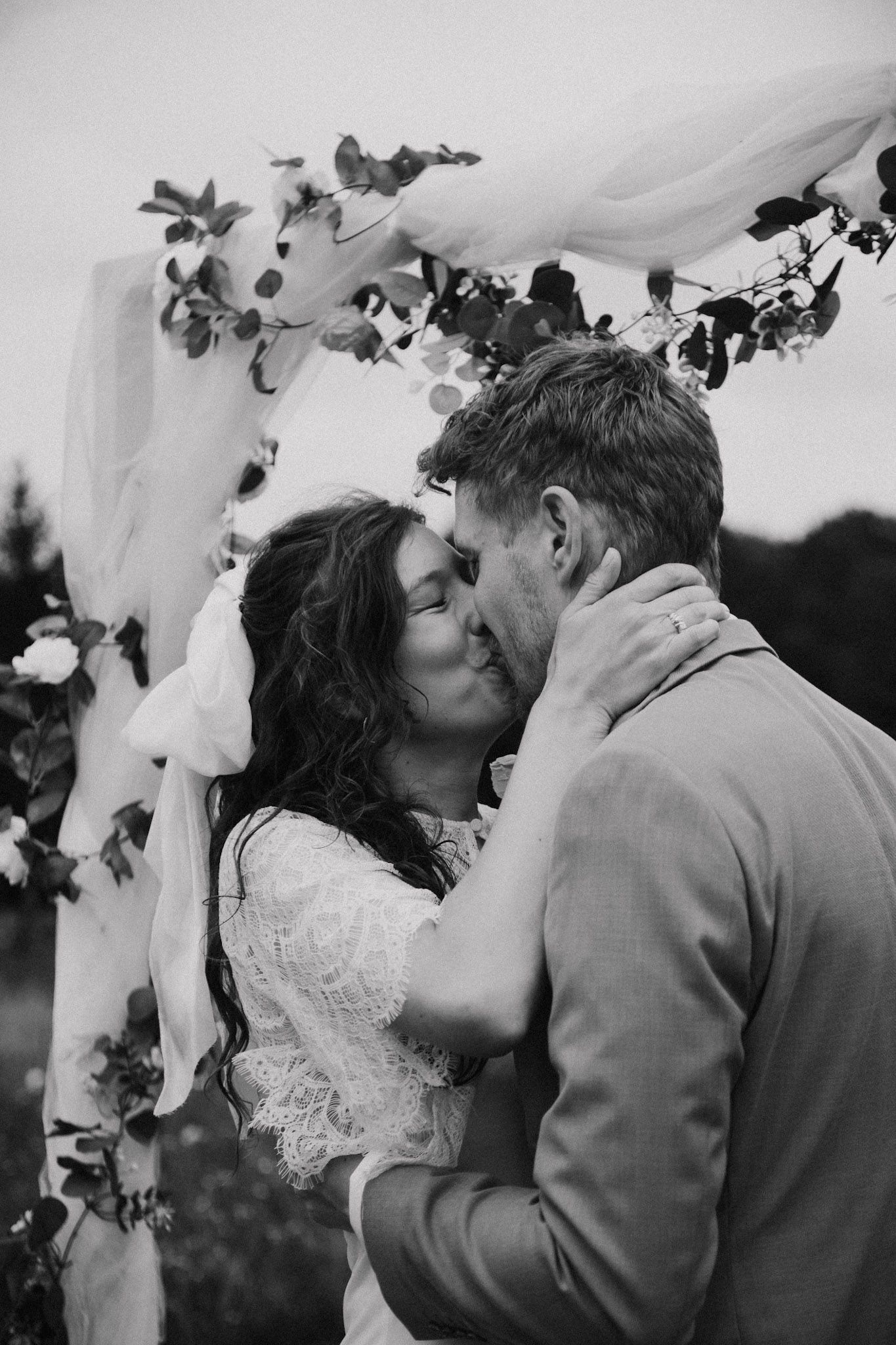 Newlyweds share a kiss under a floral wedding arch decorated with white flowers and greenery.