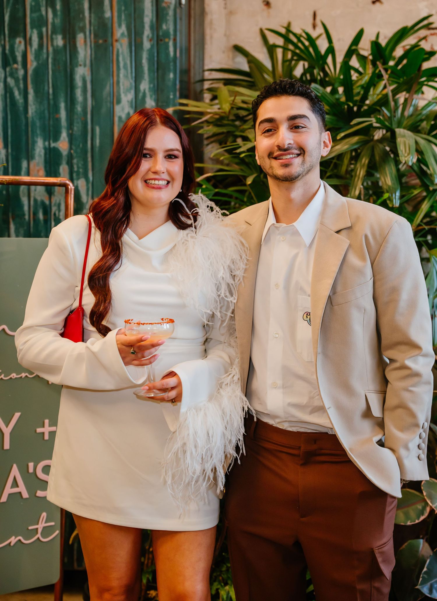 Two people wearing stylish white outfits pose together in front of greenery and a dark teal backdrop.