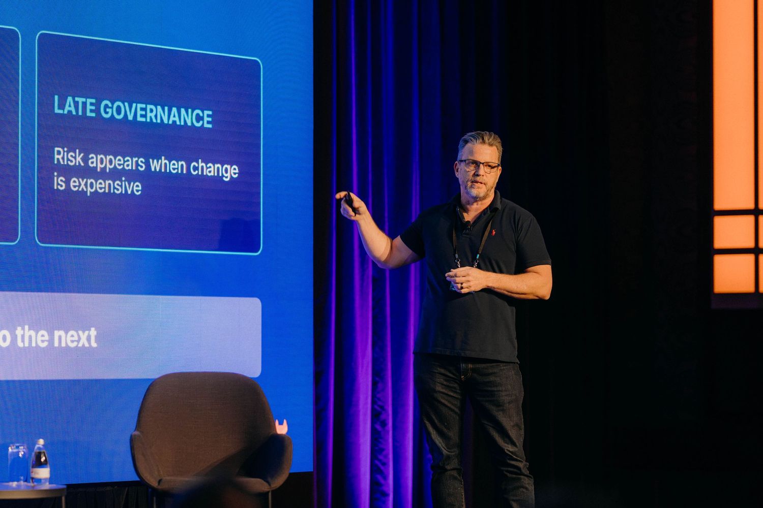 Speaker presenting slide on Late Governance at a tech conference, standing on stage with purple lighting.