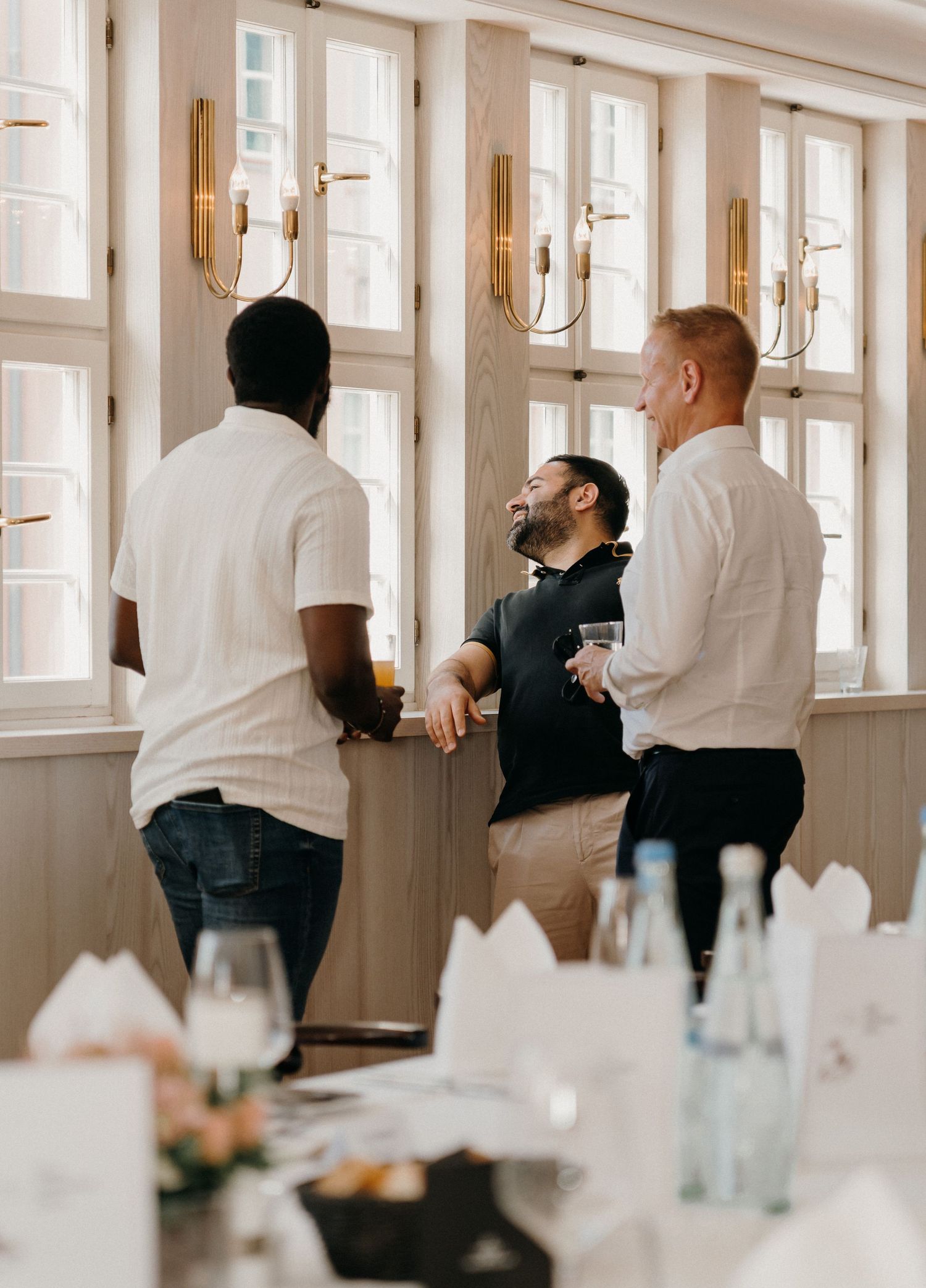 Three men laughing and socializing at an elegant restaurant event, holding drinks near large windows.