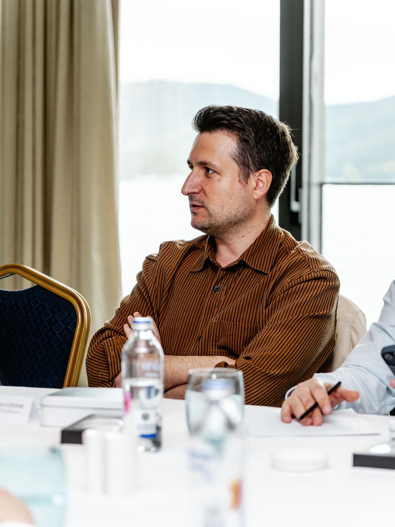 Man in brown corduroy shirt sitting at conference table listening attentively during a business meeting.