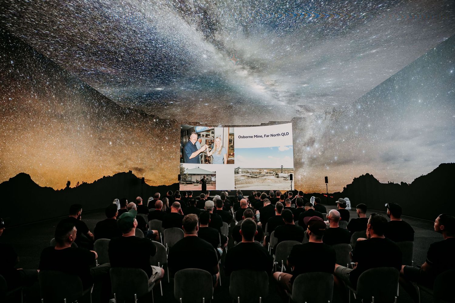 A dramatic night sky with stars and clouds stretches over a silhouetted outdoor movie theater screen and audience.