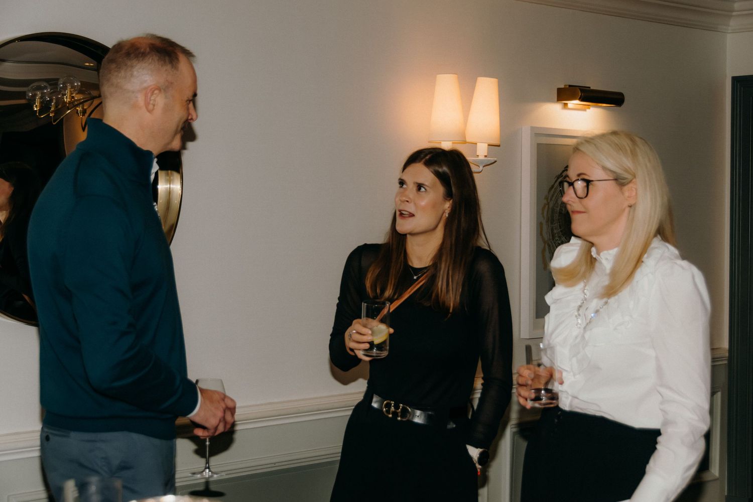 Three people engaged in casual conversation while holding drinks at an indoor social gathering.