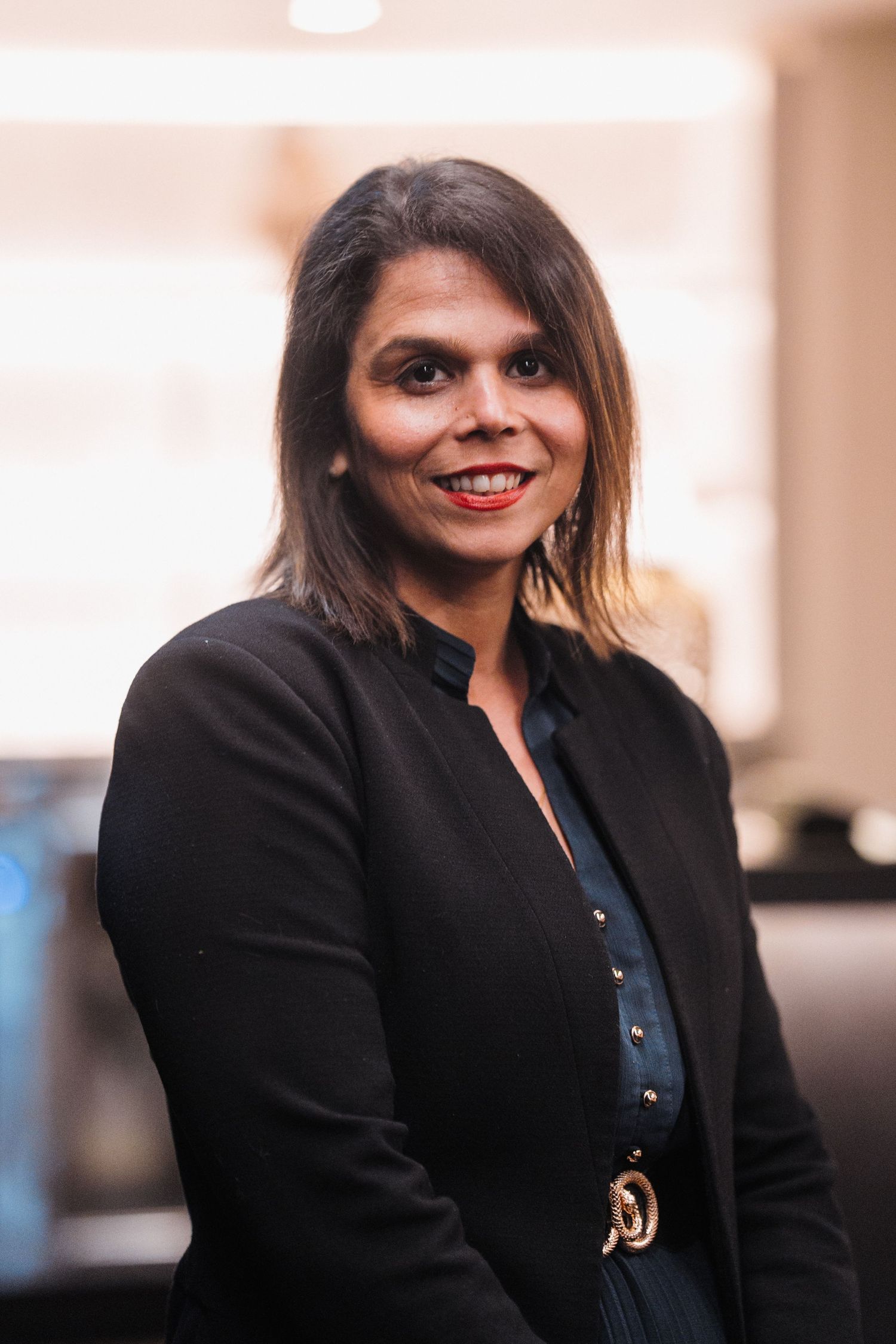 Professional wearing a black blazer and blue button-up top smiles warmly in a business setting.
