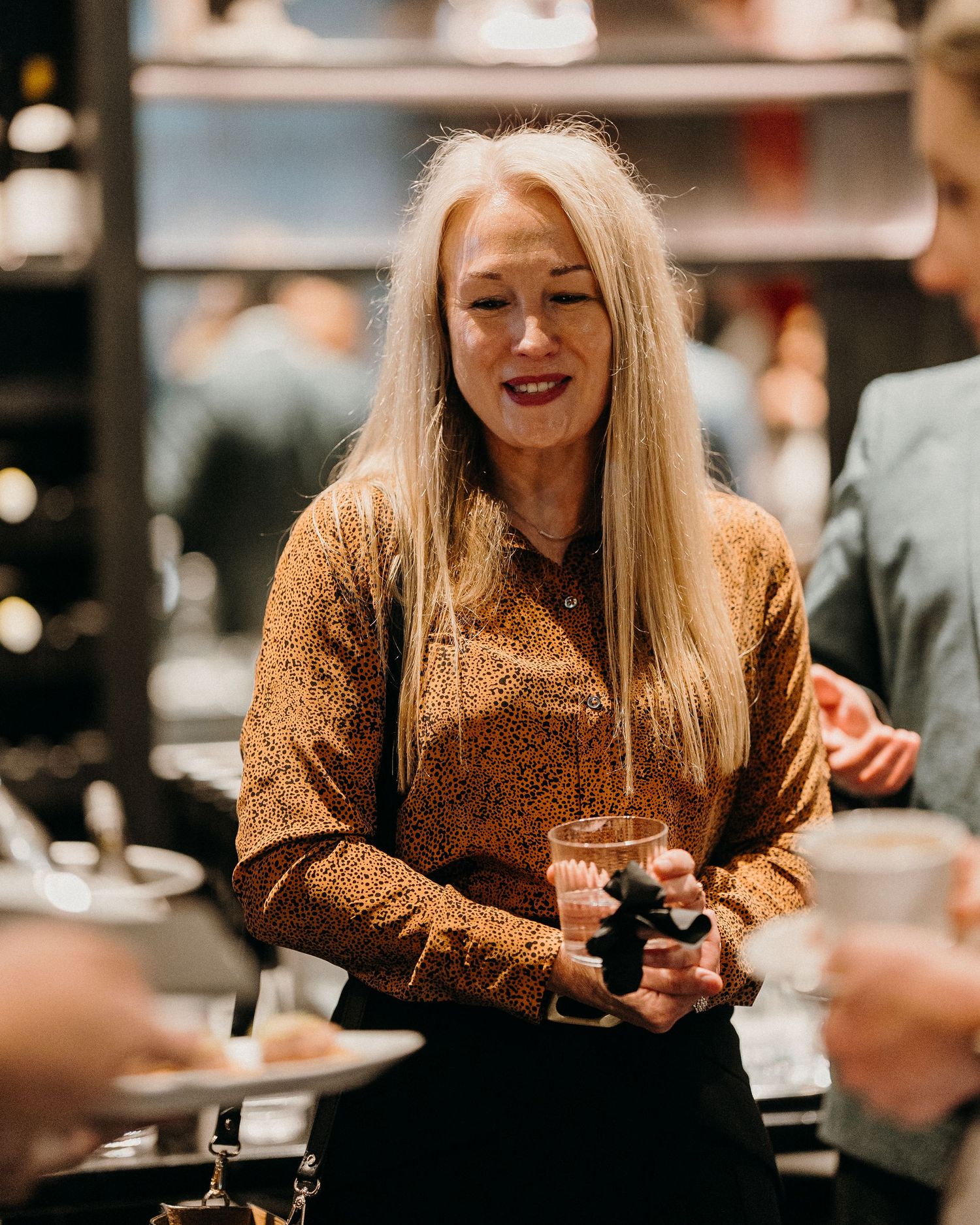 A person wearing a sparkly gold top smiles while holding a drink at a dimly lit social gathering.