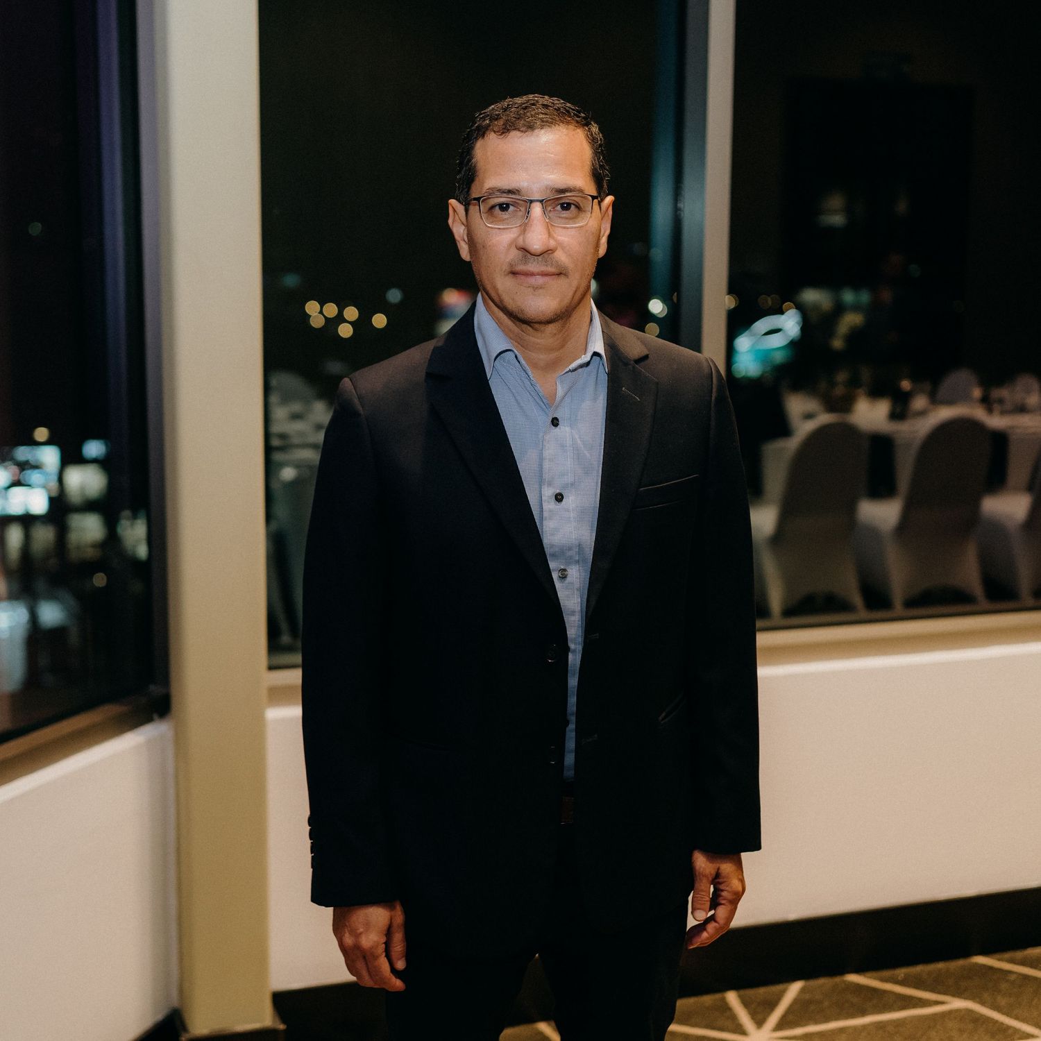 Professional wearing a dark blazer and glasses stands in an office setting with windows and lighting in the background.