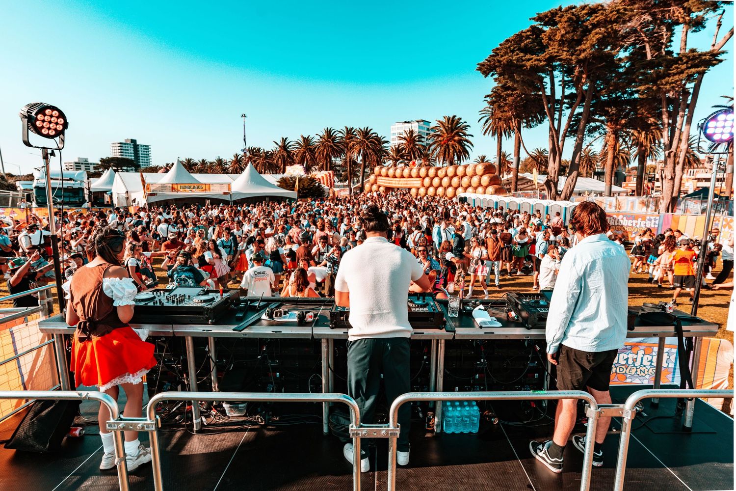Large crowd enjoying an outdoor music festival with palm trees and stage setup visible in warm sunset lighting.