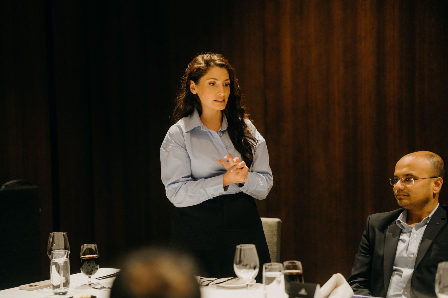 Woman in blue blouse speaking at formal dinner event with seated guests and elegant table settings.
