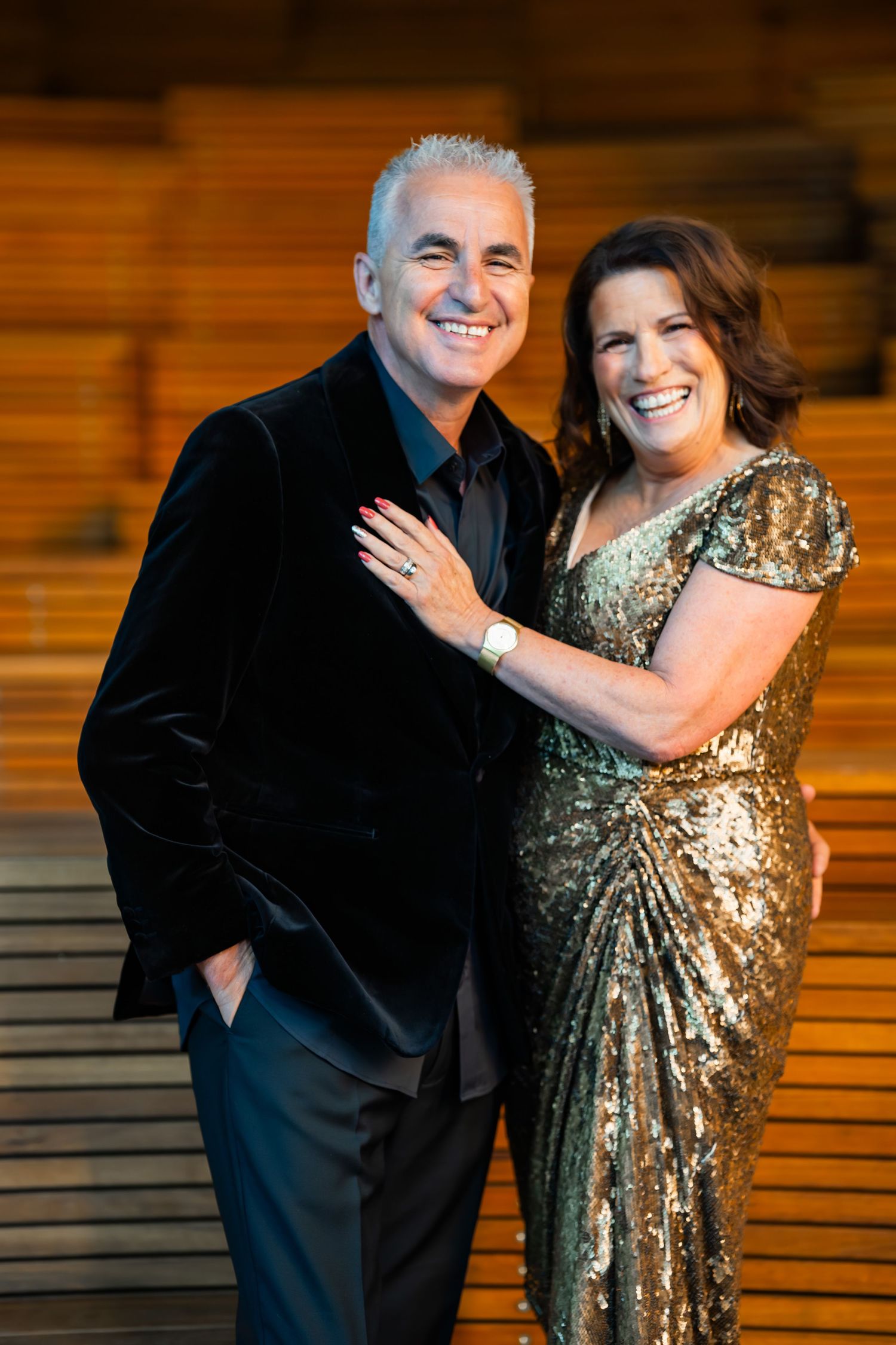 A couple embraces and laughs together, with one person wearing a sparkling gold dress and the other in a black suit.