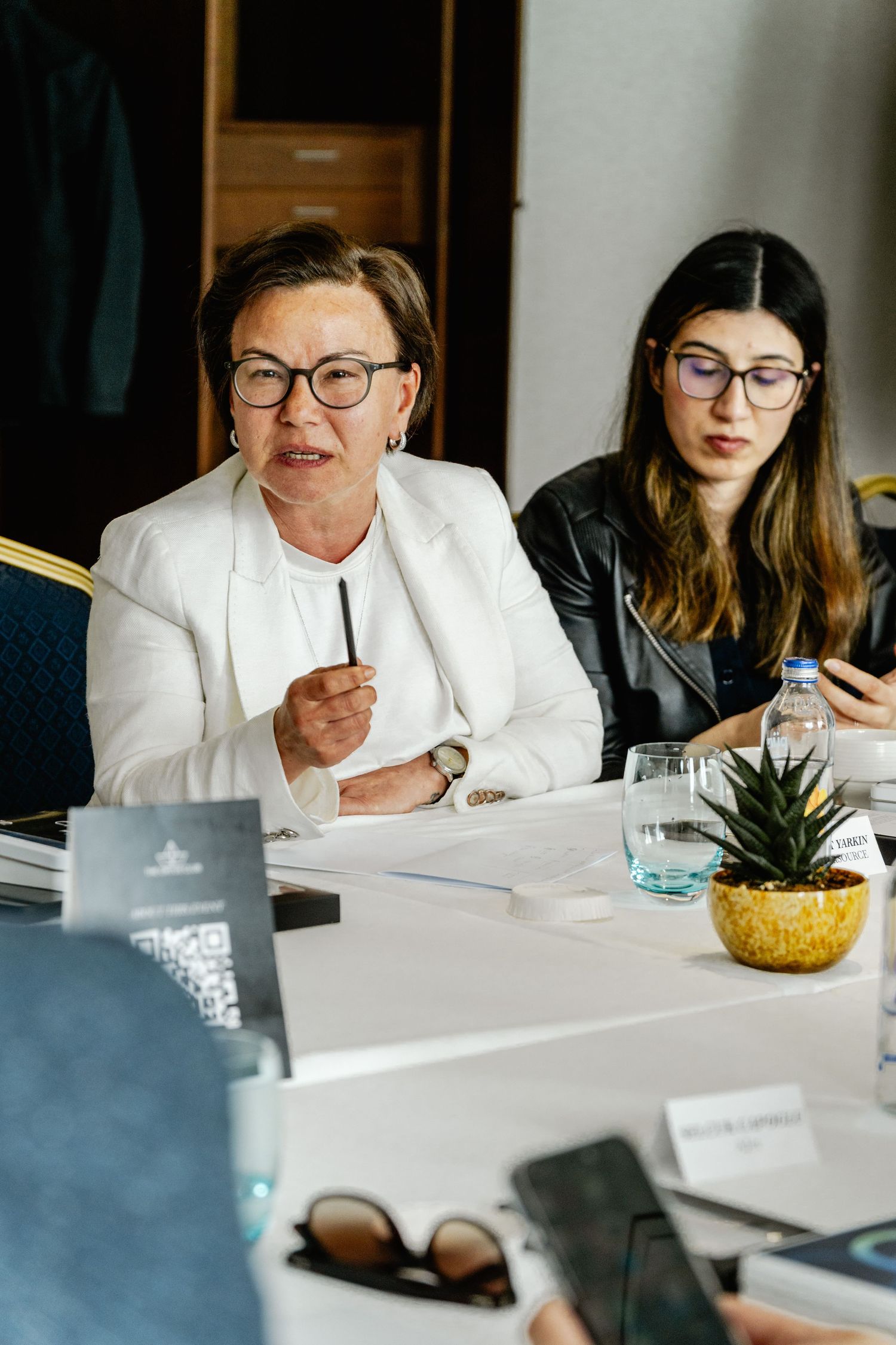 Two women wearing glasses sit at a conference table, one in a white blazer speaking during a professional meeting.