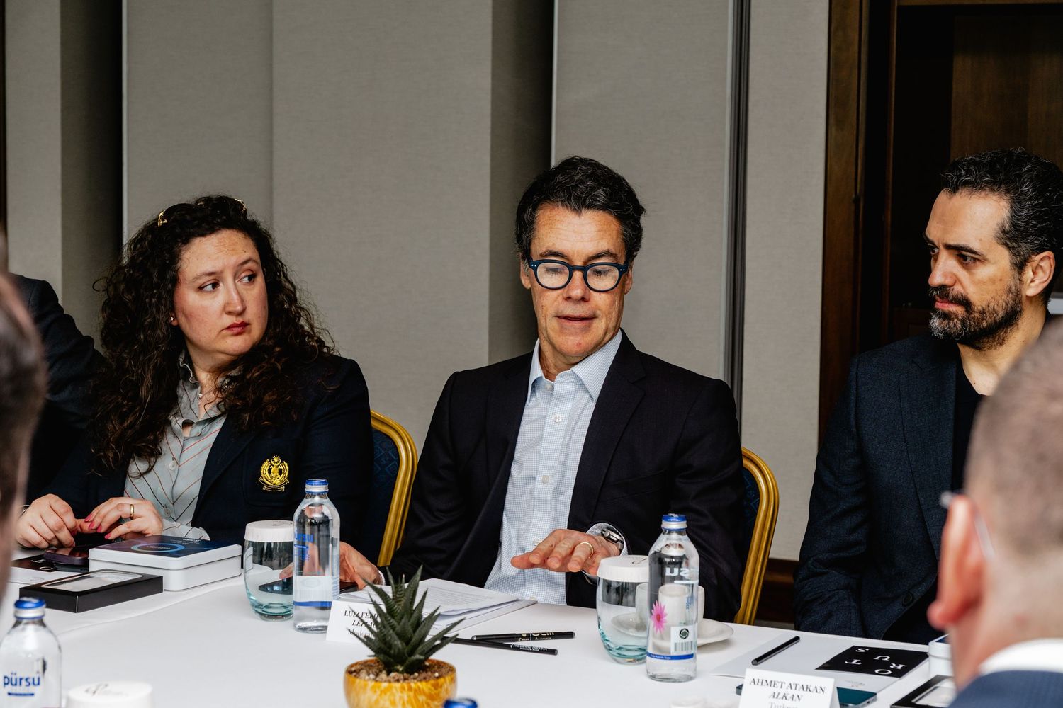 Three professionals in business attire engaged in discussion at a conference table with water bottles and a pineapple centerpiece.