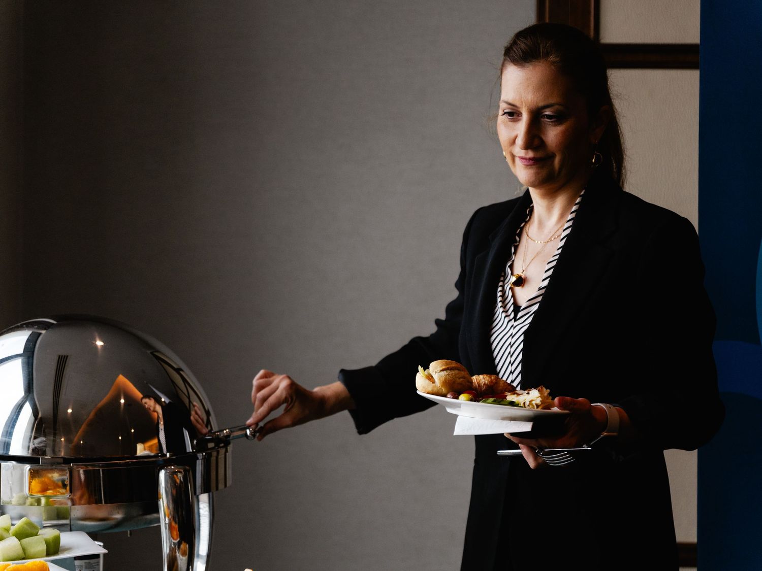 A woman in a black blazer serves food at a buffet table, holding a plate of appetizers in an elegant setting.