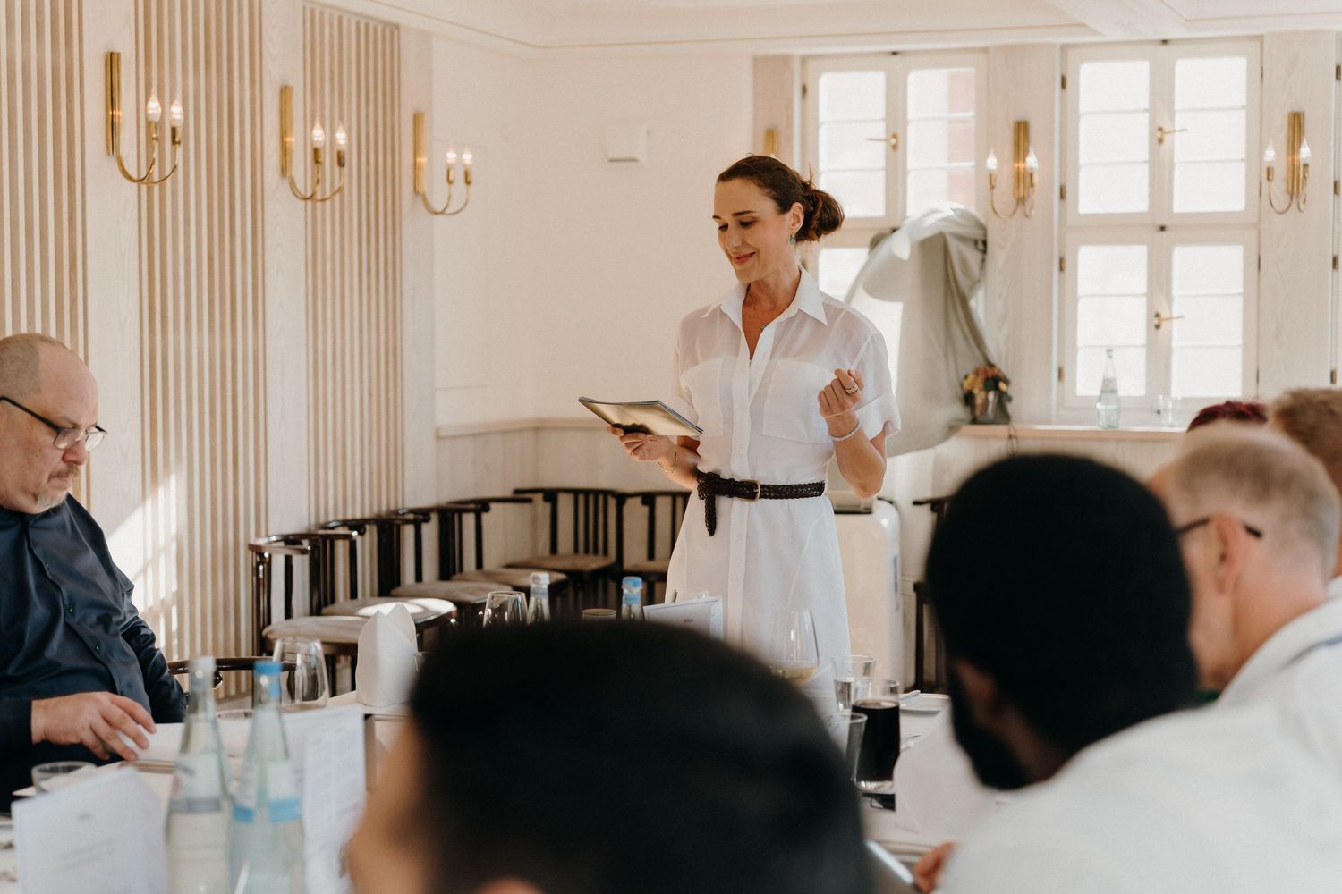 Woman in white dress presenting to a group of colleagues in an elegant, bright meeting room.
