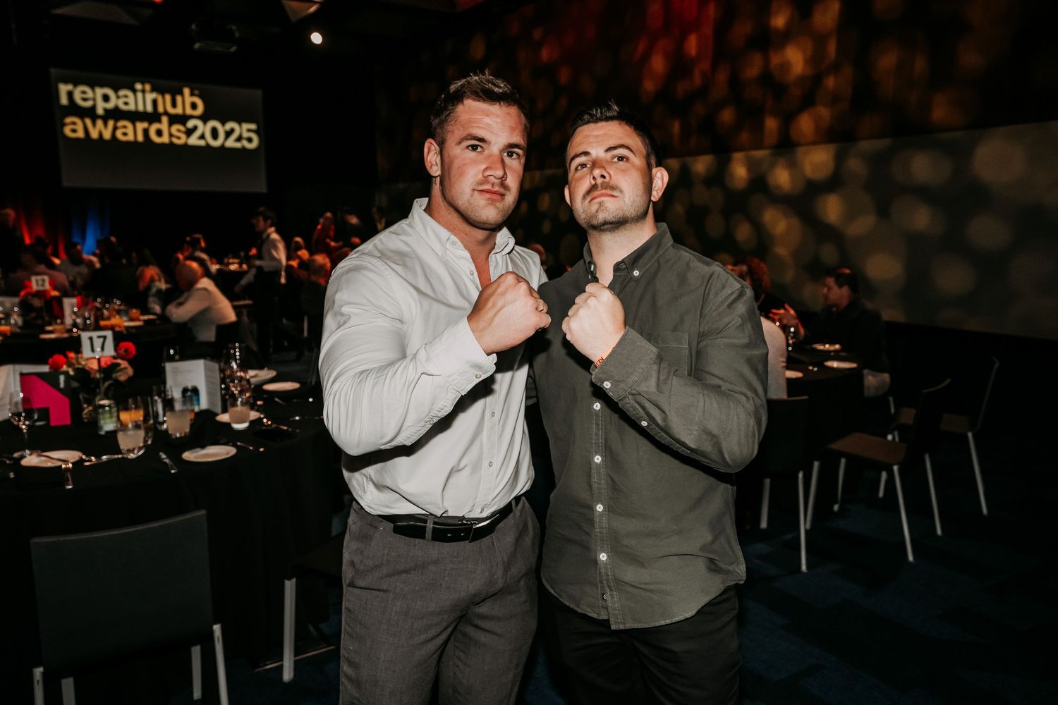 Two people pose together in formal attire at what appears to be an event or gathering with dark lighting.
