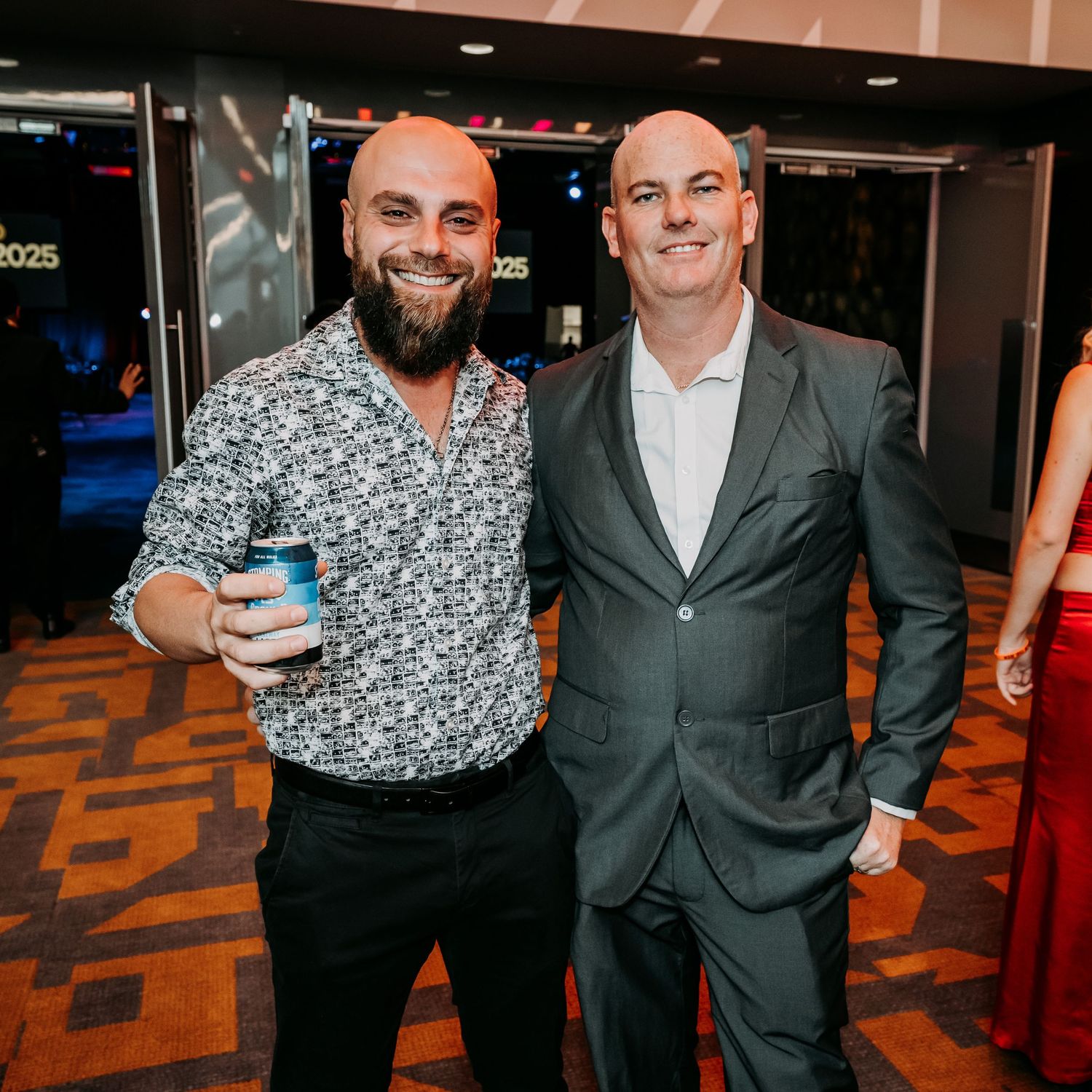 Two men in formal attire stand together at an indoor event with patterned carpet and lighting.