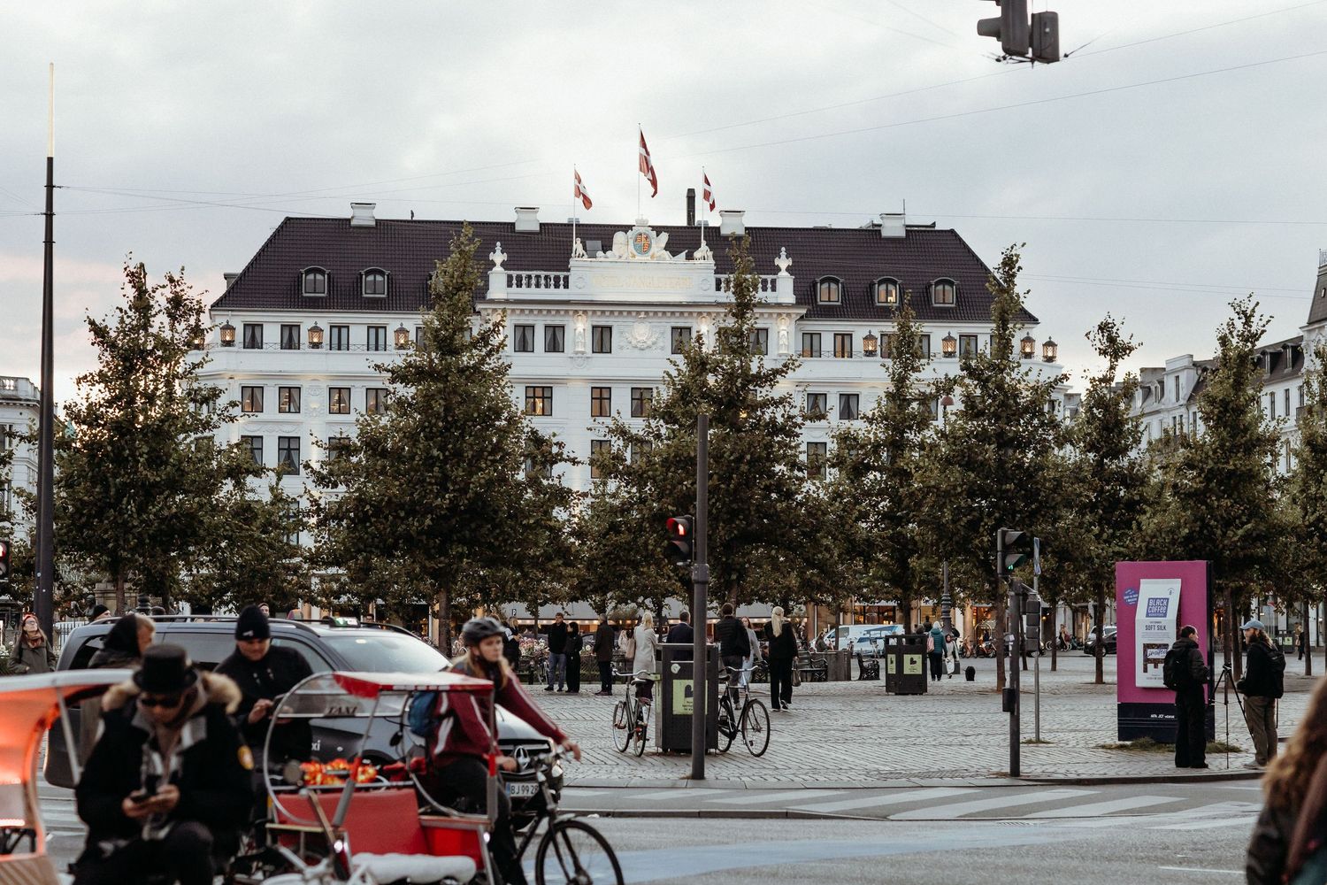 A panoramic view of a town square with white buildings, parked cars, and trees in a European setting.