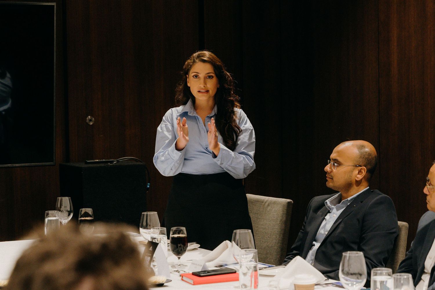 A woman in a blue shirt presents at a business dinner meeting while colleagues listen attentively at a set table.
