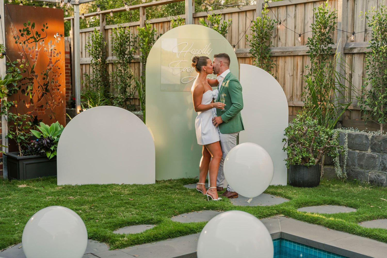 Modern backyard party setup featuring white arch decorations and glowing orb lights near a pool with stone pavers and greenery.