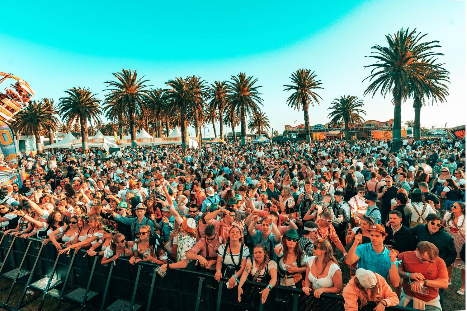 Large crowd of festival attendees gathered in front of outdoor stage with palm trees under bright turquoise sky.