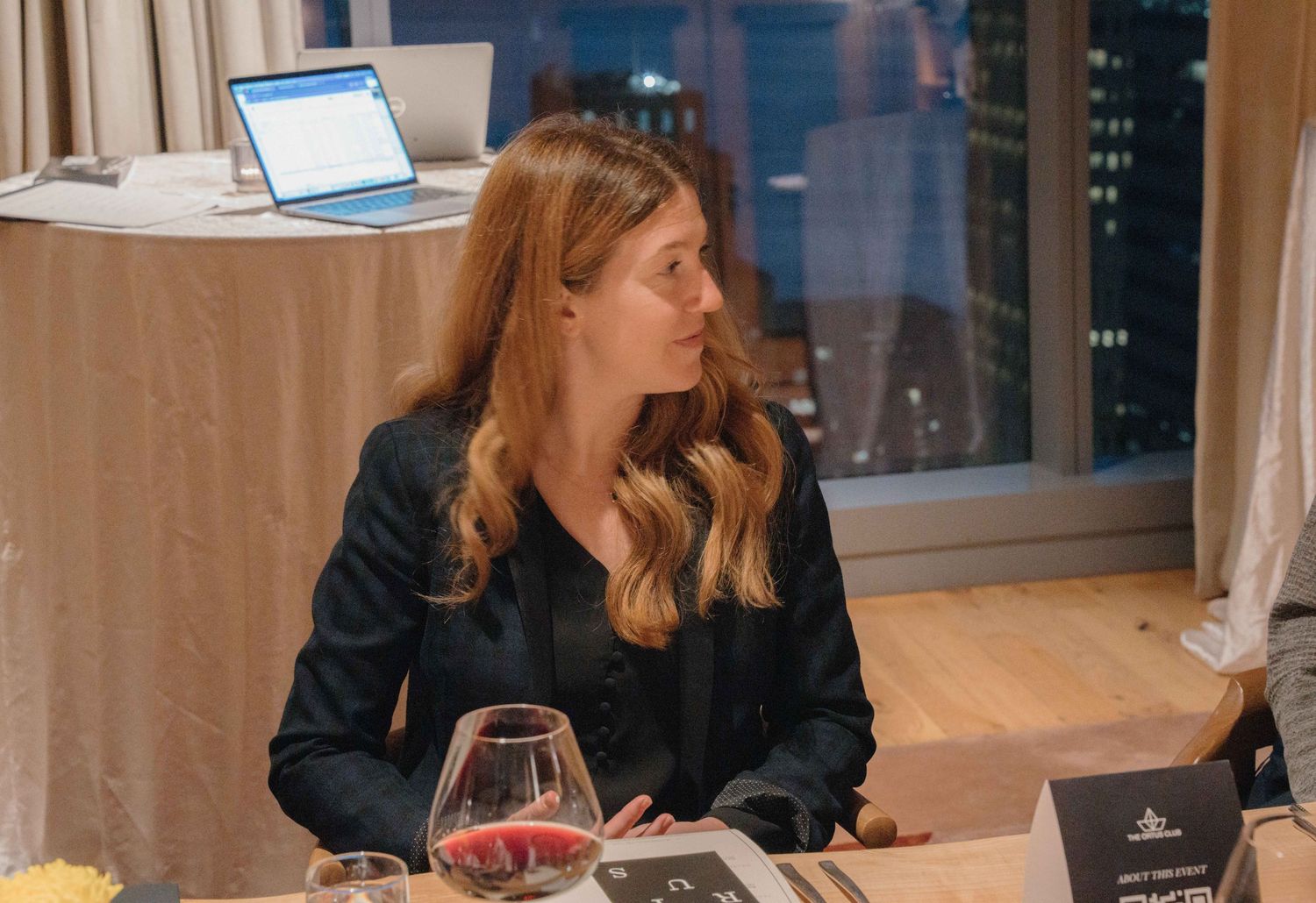 Professional working at desk in evening with laptop and wine glass in modern office setting.
