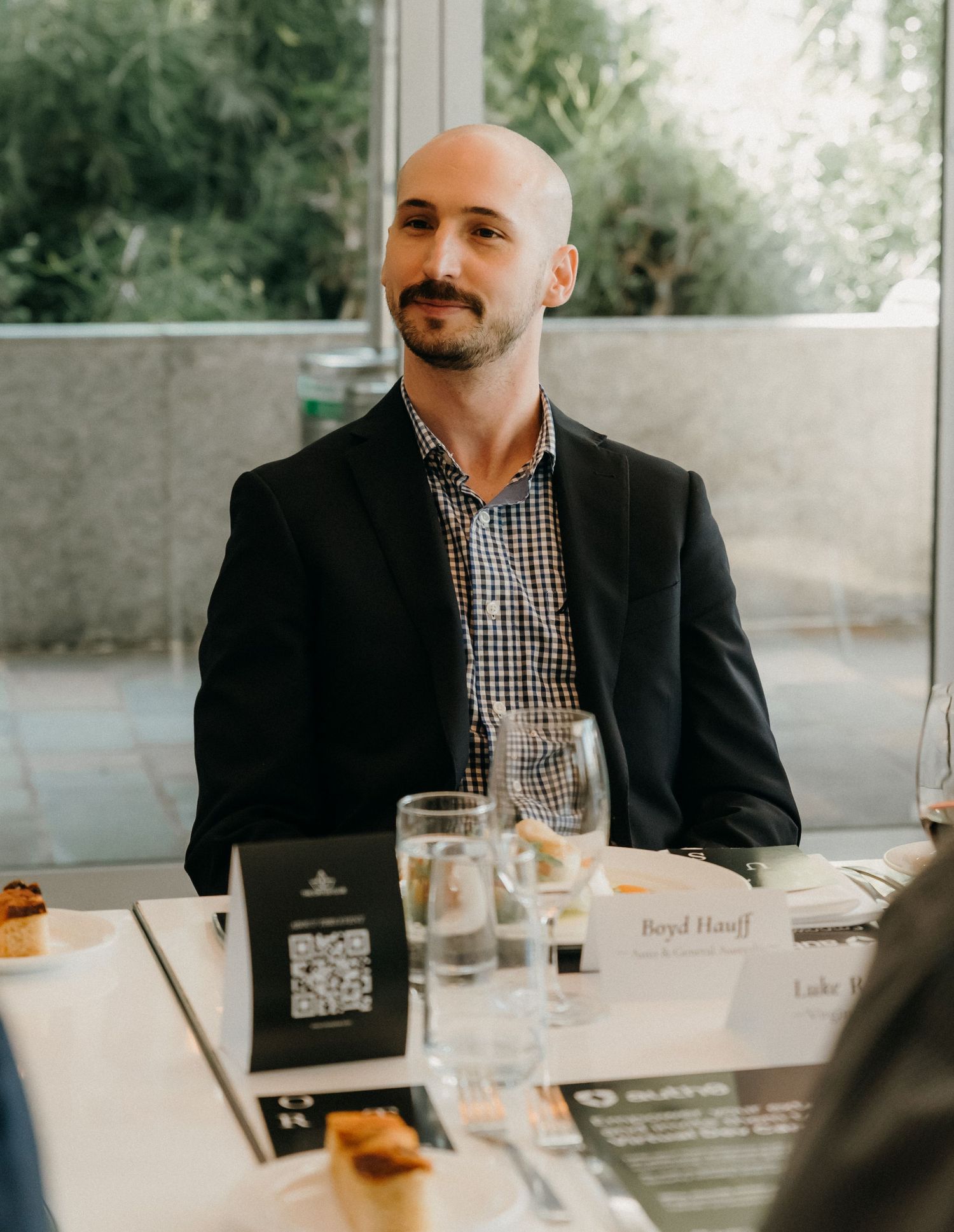 A person in a black blazer and checkered shirt sits at a restaurant table with glasses and drinks.