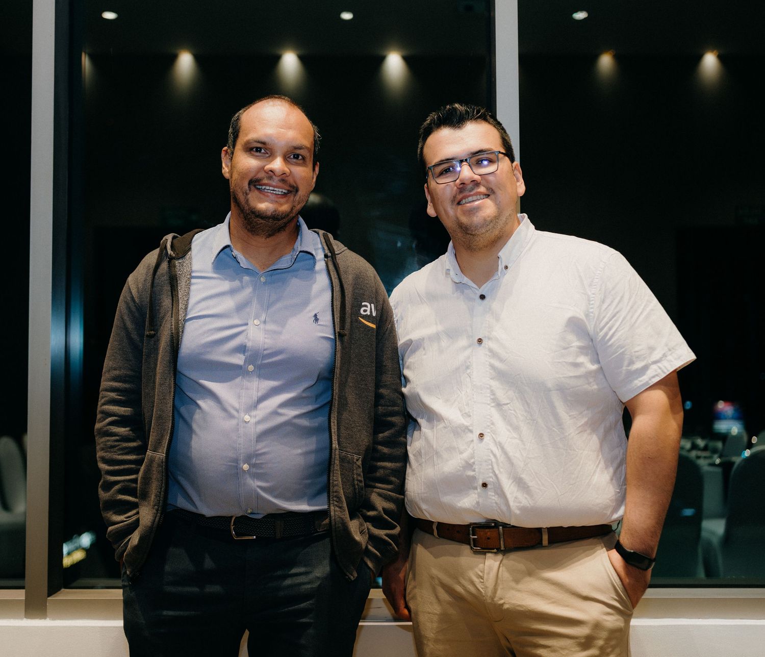 Two business professionals stand together smiling at the camera in an office setting.