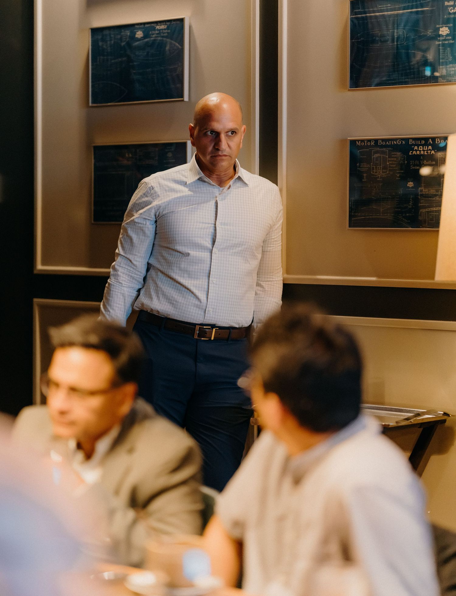 A professional in a light blue dress shirt stands in an office setting with framed documents on the wall behind them.