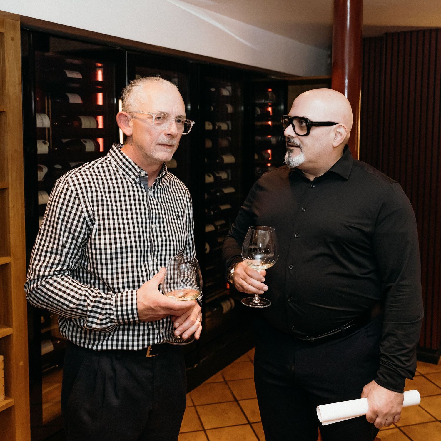 Two men in black and checkered shirts having a conversation while holding wine glasses at an indoor event.