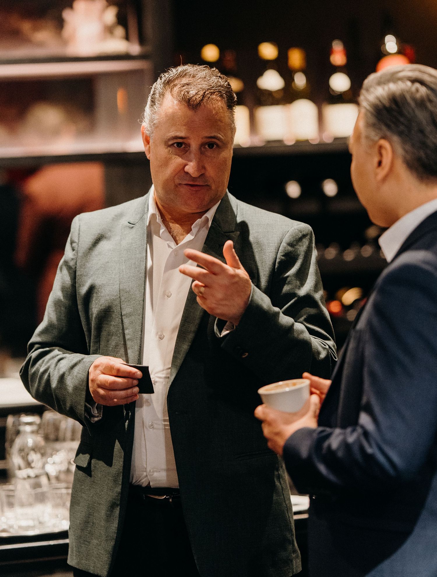 Two businessmen having a conversation over drinks in an upscale bar with wine bottles in the background.