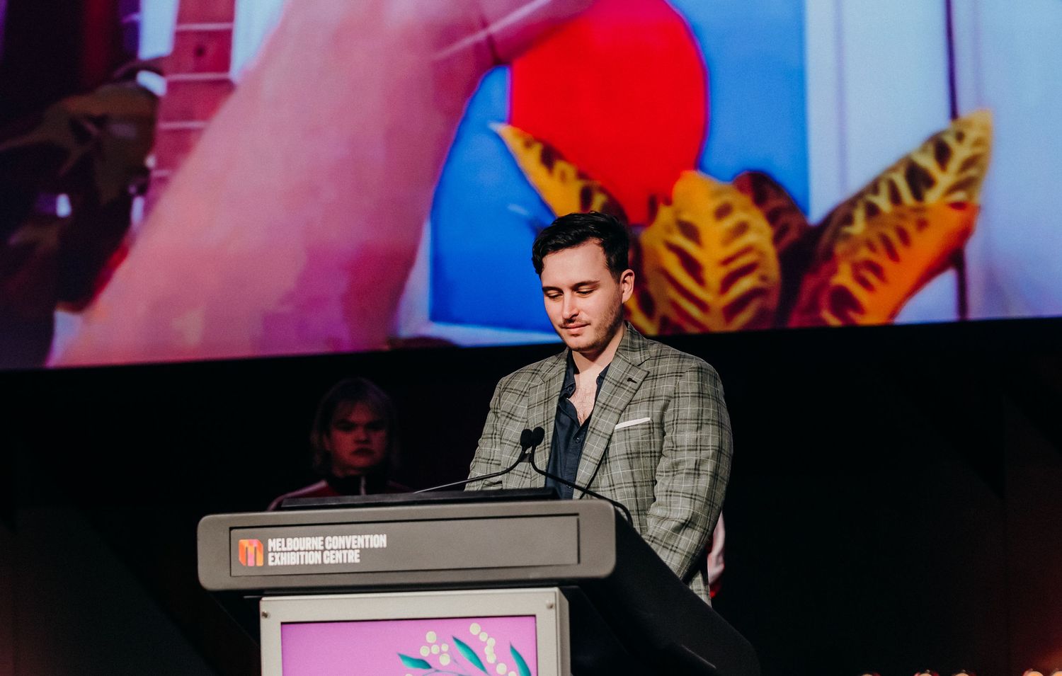 A presenter stands at a podium speaking to an audience during a conference presentation.