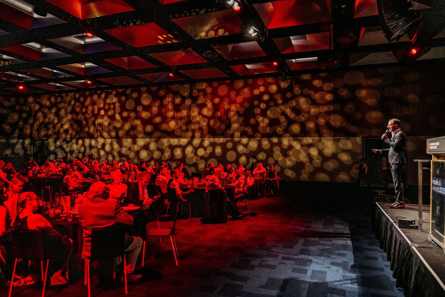 A concert venue illuminated in red lighting with silhouettes of audience members visible in the background.